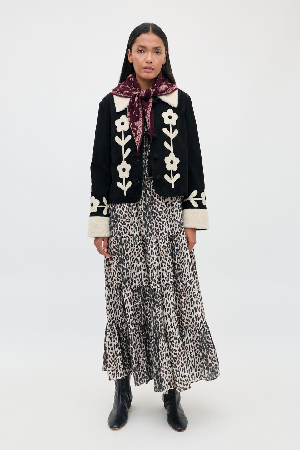 A woman stands against a plain white background wearing the Gabriel - Nina Leopard Mocha dress, styled with a black jacket featuring white floral designs, black boots, and a maroon scarf tied around her neck.
