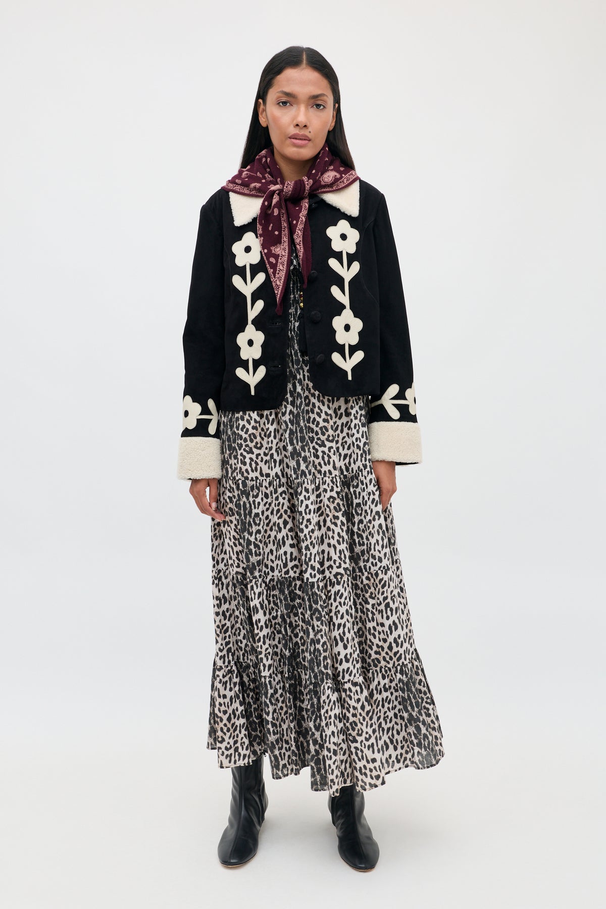 A woman stands against a plain white background wearing the Gabriel - Nina Leopard Mocha dress, styled with a black jacket featuring white floral designs, black boots, and a maroon scarf tied around her neck.