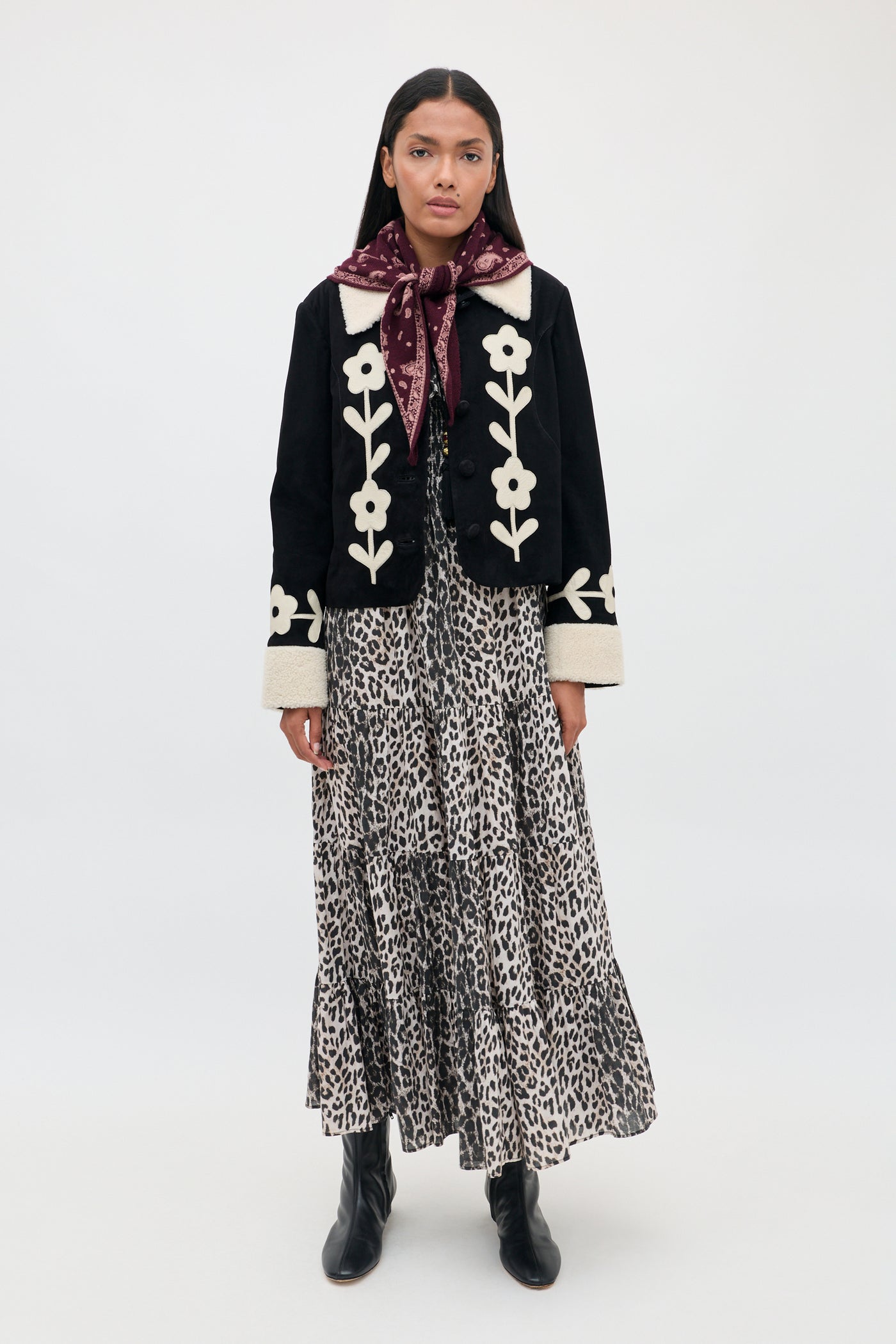 A woman stands against a plain white background wearing the Gabriel - Nina Leopard Mocha dress, styled with a black jacket featuring white floral designs, black boots, and a maroon scarf tied around her neck.