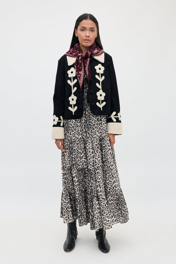 A woman stands against a plain white background wearing the Gabriel - Nina Leopard Mocha dress, styled with a black jacket featuring white floral designs, black boots, and a maroon scarf tied around her neck.