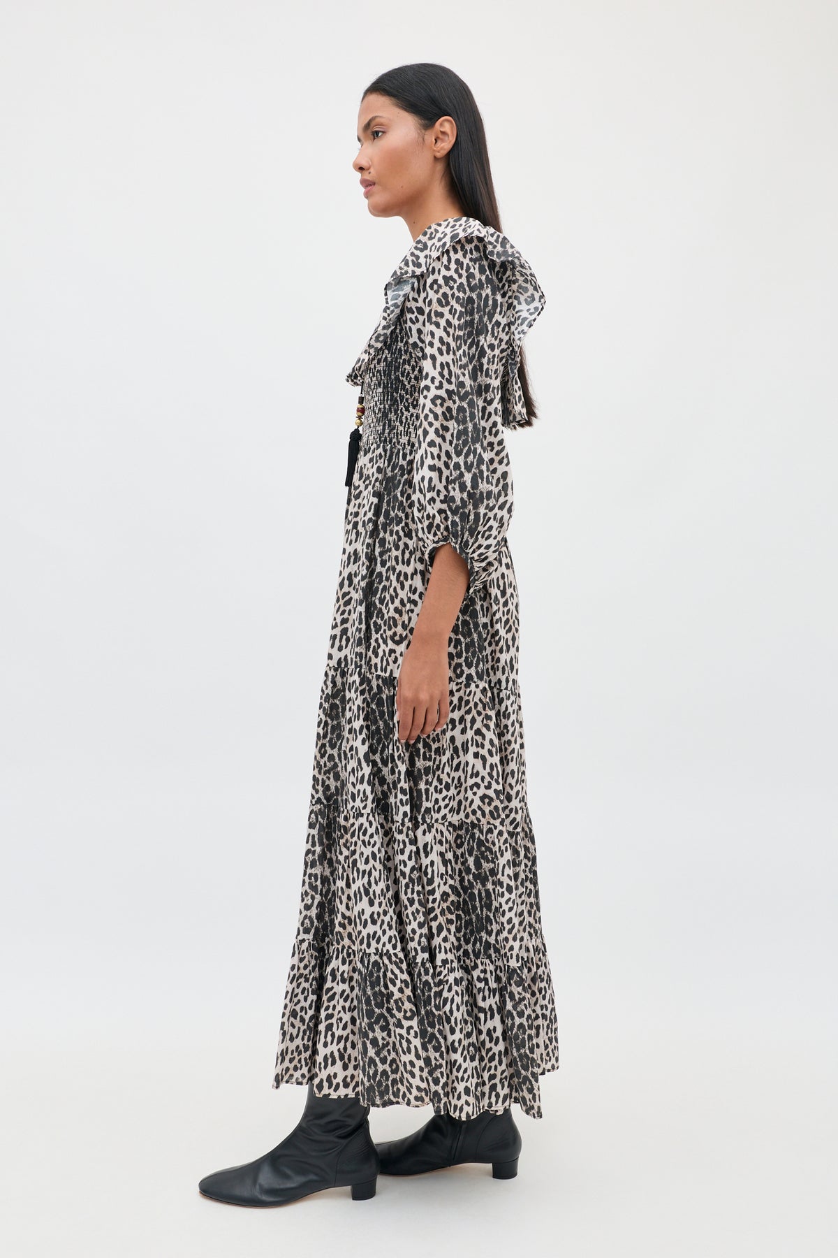 A woman stands in profile wearing the Gabriel - Nina Leopard Mocha dress, a long, loose-fitting design with long sleeves and a leopard-print pattern, paired with black ankle boots. The background is plain and light-colored.