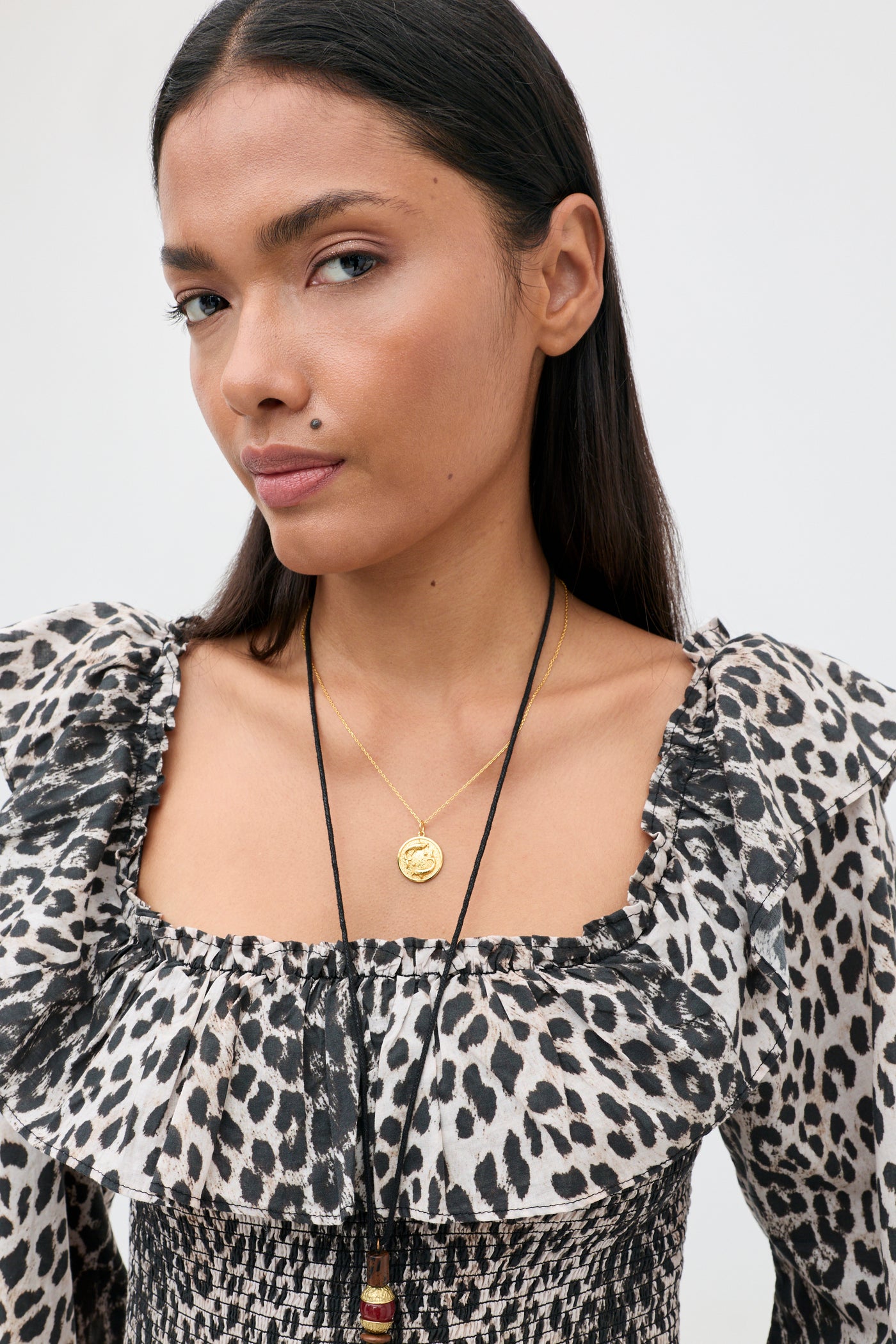A person with long dark hair wears the Gabriel - Nina Leopard Mocha top, which features ruffled details, along with two necklaces—one gold and one black. They have a neutral expression and a visible beauty mark above their lip.
