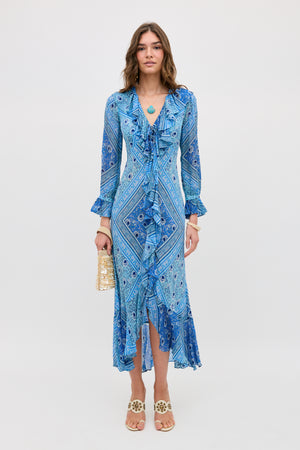 A woman stands facing forward in a Gracye - Boho Scarf Blue dress with long sleeves and a V-neckline, holding a beige handbag and wearing white decorative sandals. The background is plain white.