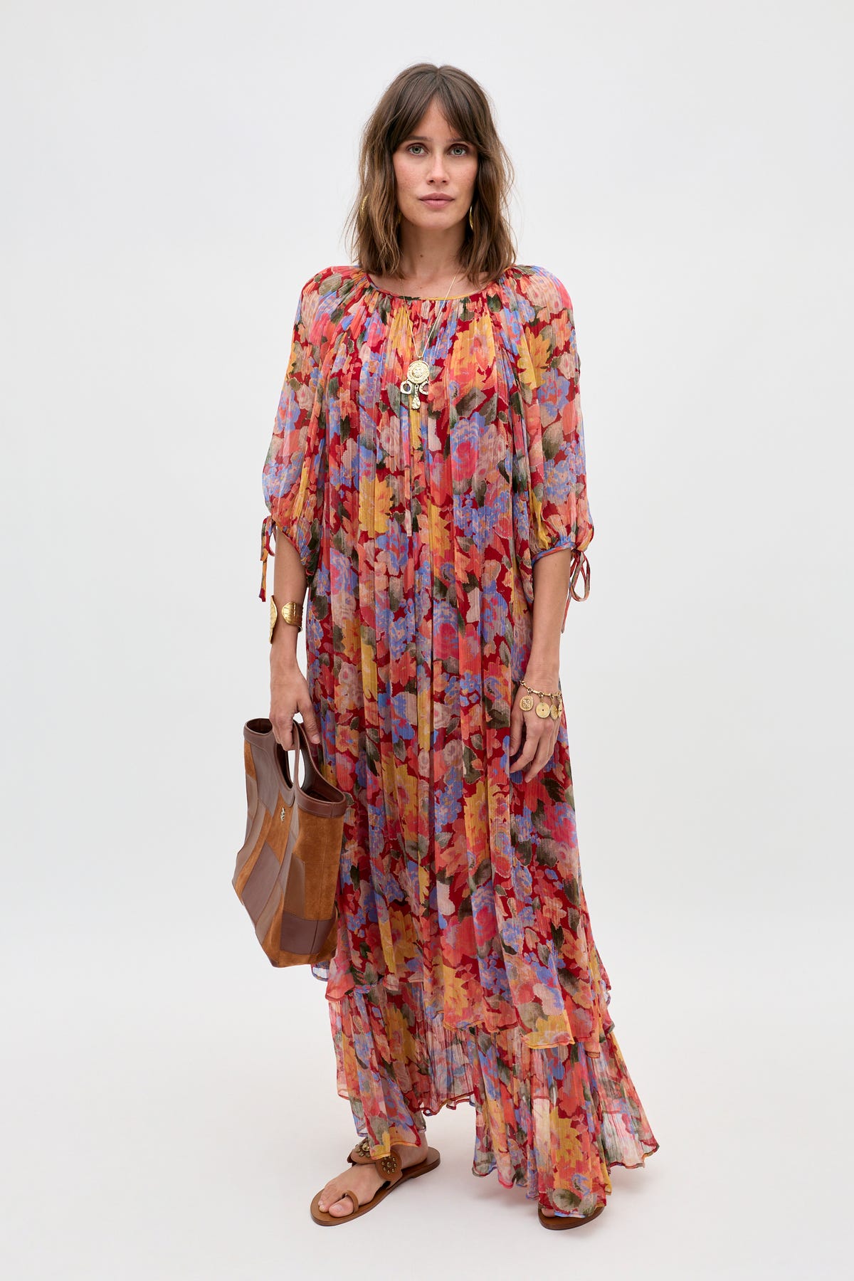 A woman wearing the Hayden - Chelsea Floral Red long, colorful floral dress with a ruffled hem, paired with brown sandals and gold jewelry, holds a brown tote bag while standing against a plain light background.