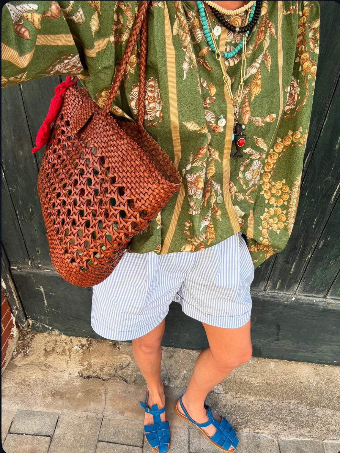 A person wearing a green patterned shirt, striped shorts, blue sandals, and layered necklaces carries the Lucine - Tan leather woven bag by Dragon Diffusion while standing on stone pavement before an aged green wooden door.