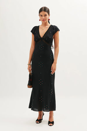 A woman poses against a white background wearing the Iggy - Rose Trail Embroidery Black, a versatile floor-length dress with a deep V-neck, cap sleeves, and subtle floral embroidery. She accessorizes with a small brown handbag and black heels.