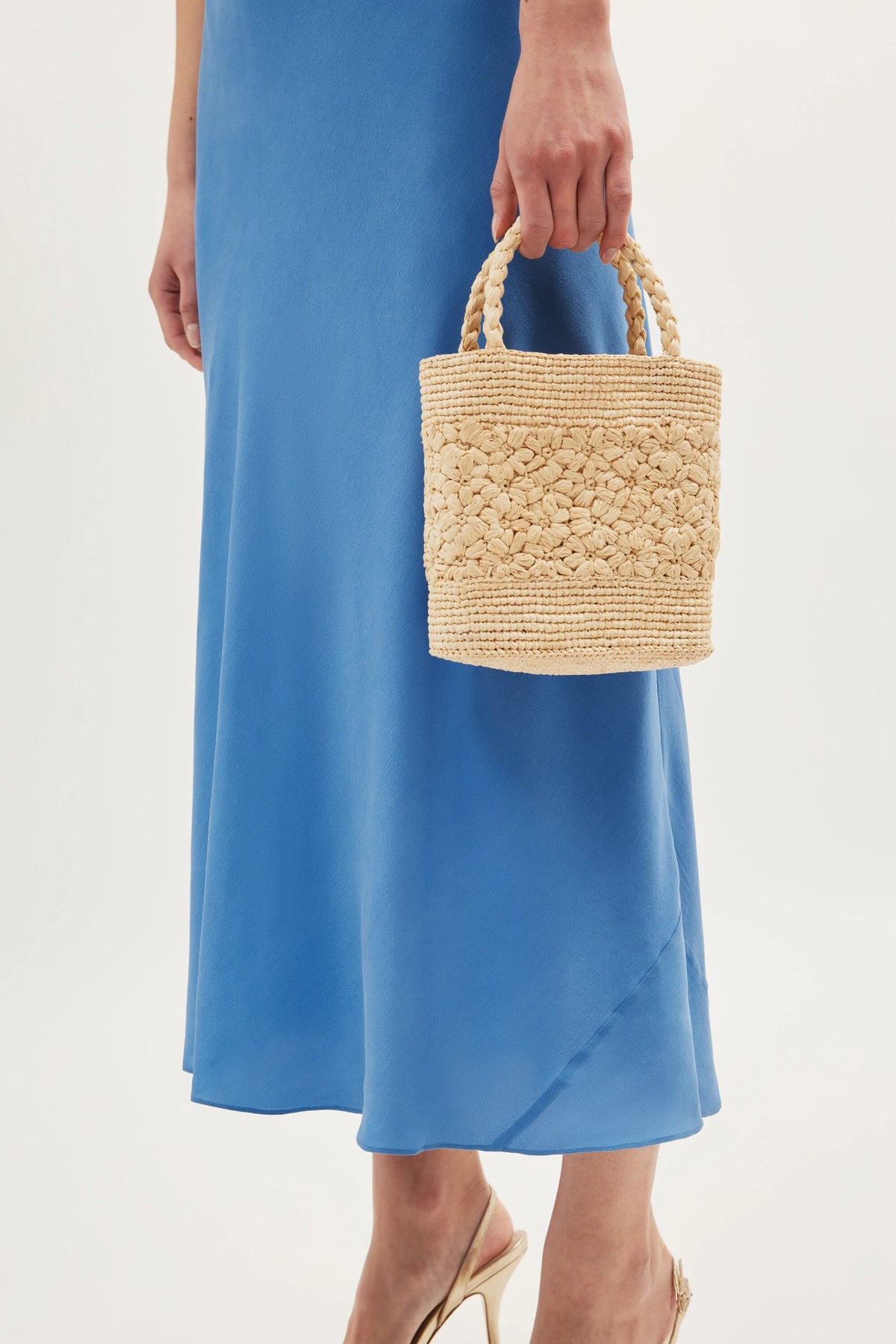 A person in the Ilaria - Blue silk skirt and heeled shoes holds a small woven straw handbag with two short handles, set against a minimal white background.