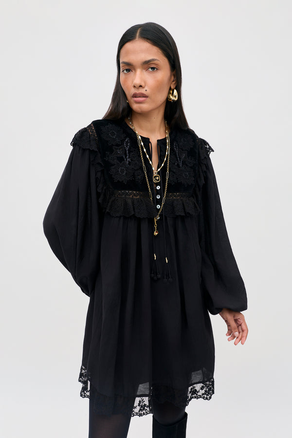 A woman stands against a plain light background wearing the Inez - Lilly Embroidery Black dress, a loose black style featuring lace details on the collar and hem. She completes her look with layered necklaces, gold earrings, and wears her long dark hair down.