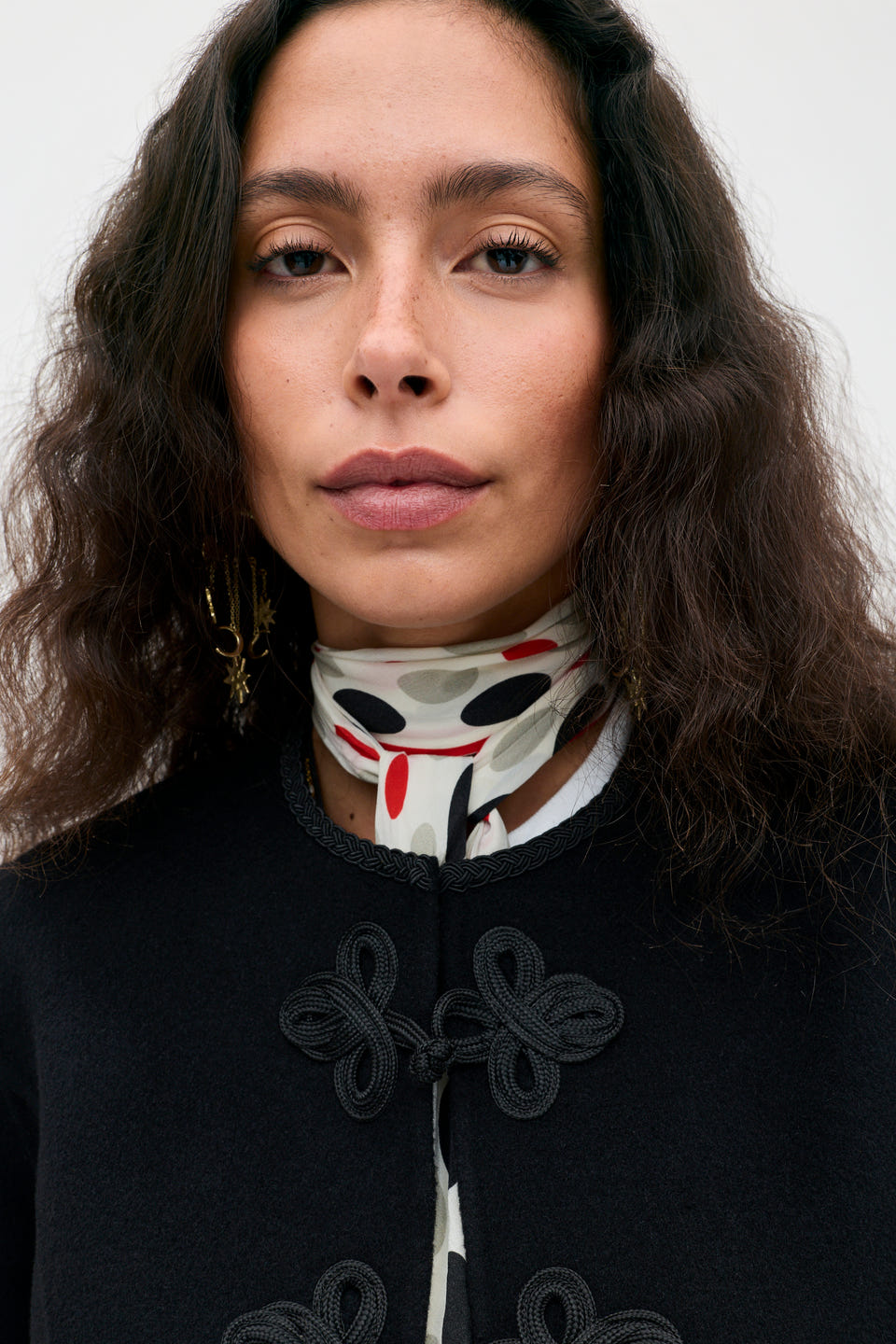 Woman with wavy dark hair, wearing the Inga - Black jacket with decorative knots, a white scarf with black and red dots, and gold earrings, looking directly at the camera against a plain background.