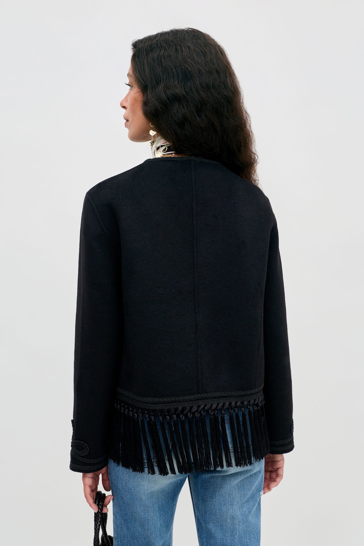 A woman stands with her back to the camera, wearing the Inga - Black jacket with fringe details along the bottom hem, paired with blue jeans and holding a small black item. Her hair is loose and wavy, and the background is plain light gray.