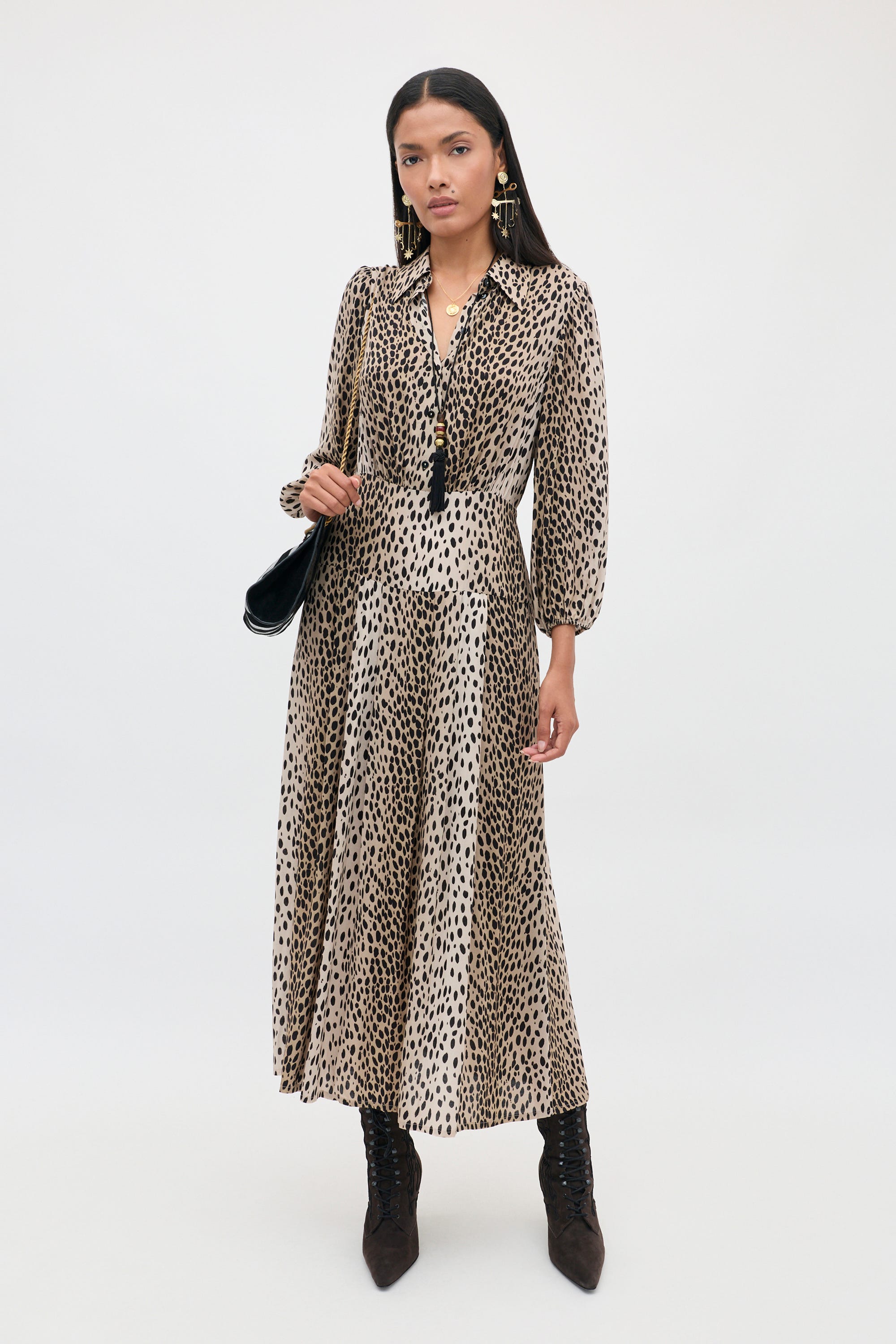 A woman stands against a plain white background, wearing the Izzy - Antelope Spot Black long-sleeved shirt dress with elasticated cuffs, styled with black boots, earrings, a necklace, and holding a black shoulder bag.