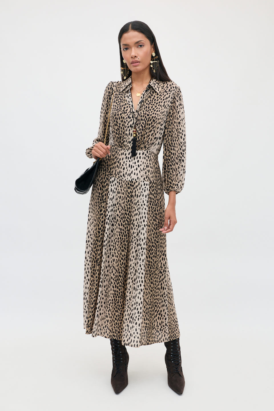 A woman stands against a plain white background, wearing the Izzy - Antelope Spot Black long-sleeved shirt dress with elasticated cuffs, styled with black boots, earrings, a necklace, and holding a black shoulder bag.