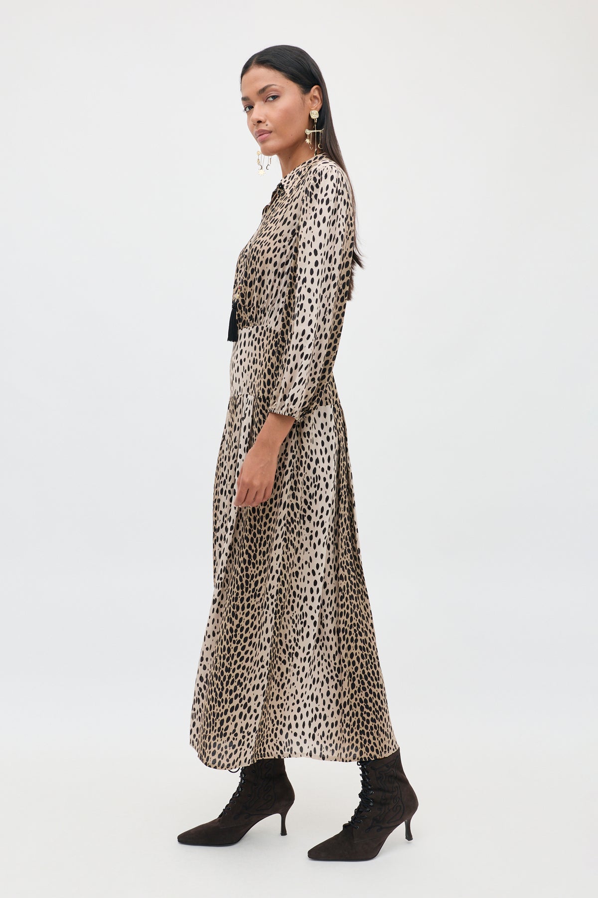 A woman stands sideways against a plain white background, wearing the Izzy - Antelope Spot Black long shirt dress with elasticated cuffs, styled with a black belt, statement earrings, and black lace-up ankle boots.
