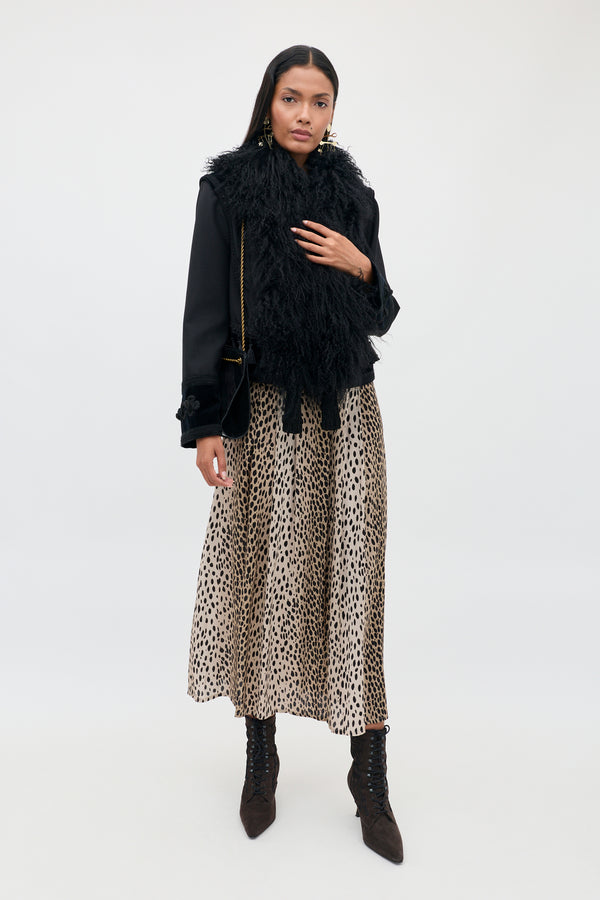 A woman with long dark hair and a neutral expression stands against a plain background wearing the “Izzy - Antelope Spot Black” leopard print pleated skirt, paired with a black fuzzy jacket featuring elasticated cuffs, lace-up black boots, and holding a black handbag with a gold chain strap.