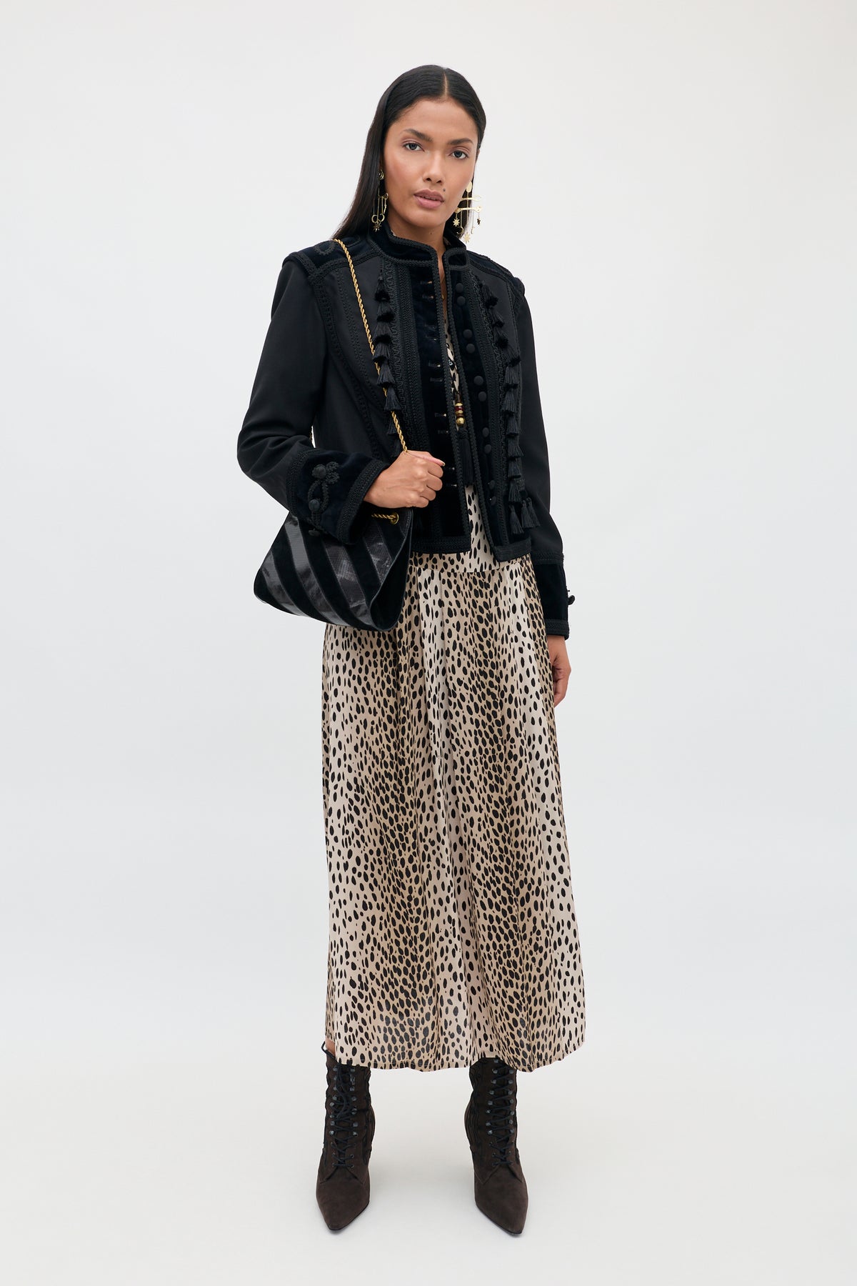 A woman with long dark hair and a neutral expression stands against a plain background, wearing the Izzy - Antelope Spot Black long leopard print skirt, paired with a black jacket featuring elasticated cuffs, black lace-up boots, and holding a black shoulder bag.
