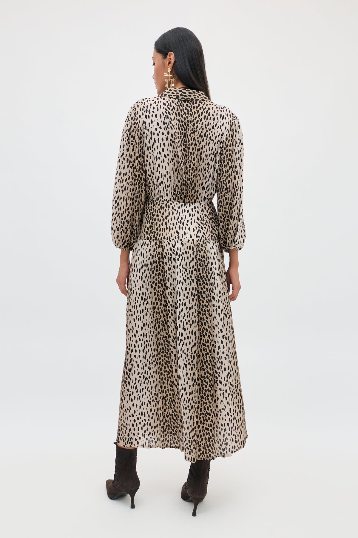 Woman with long dark hair wears the Izzy - Antelope Spot Black, a long, flowy shirt dress with elasticated cuffs and black heeled boots, standing against a plain light background, shown from the back.