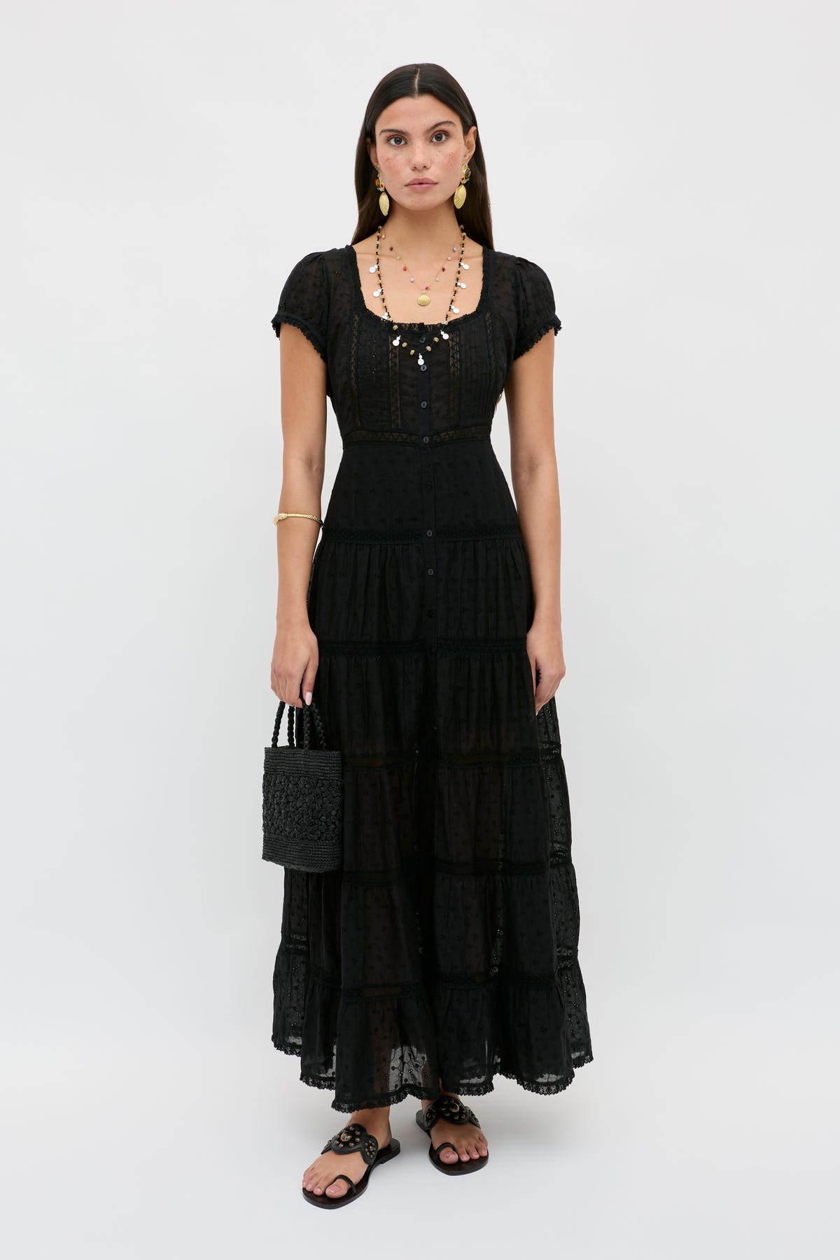 A woman with straight brown hair stands facing forward against a plain white background, wearing the Jaclyn - Black long, tiered black dress with short sleeves. She pairs it with a black woven handbag, black sandals, and gold jewelry.