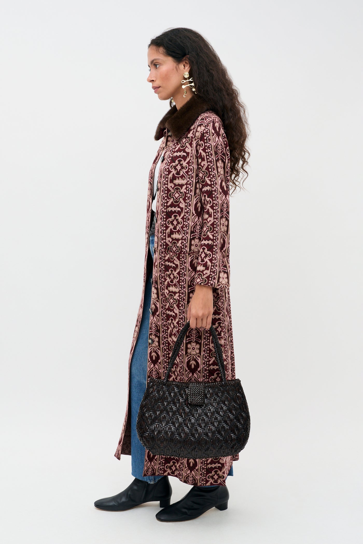 A woman stands in profile wearing the Jacoba - Tapestry Bloom Bordeaux, a long patterned coat, paired with blue jeans and black boots. She holds a black textured handbag, accessorizes with large earrings, and wears her long dark curly hair down. The background is plain white.