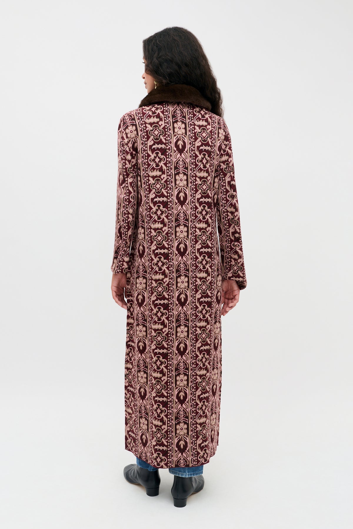 A person with long dark hair, seen from behind, stands against a plain light background wearing the Jacoba - Tapestry Bloom Bordeaux—a long, patterned maroon and beige coat with a brown collar—paired with blue jeans and black shoes.