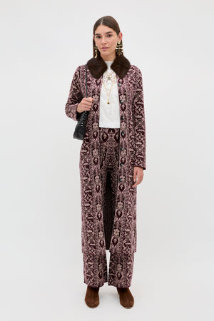 A woman wears the Ceara - Tapestry Bloom Bordeaux long patterned coat with matching pants, paired with a white top, layered necklaces, brown shoes, and a black handbag, standing against a plain white background.