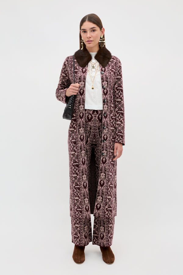 A woman wears the Ceara - Tapestry Bloom Bordeaux long patterned coat with matching pants, paired with a white top, layered necklaces, brown shoes, and a black handbag, standing against a plain white background.
