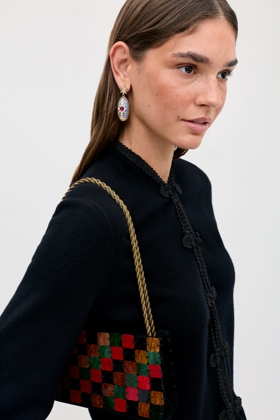 A woman wears a black top and a Gracelyn - Multi handbag featuring a multicolored checkerboard pattern and a gold chain strap.