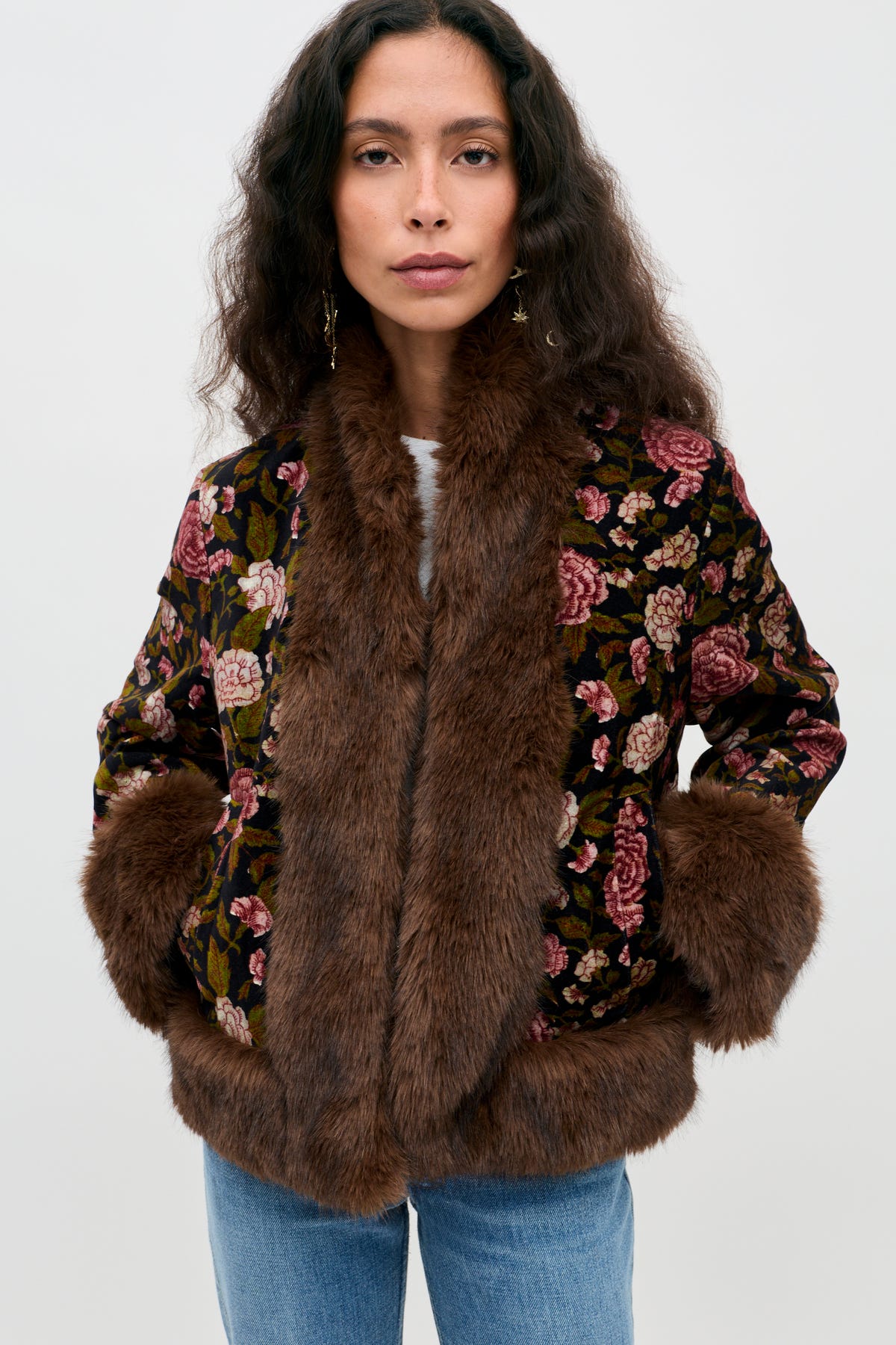 A woman wearing the Jorgia - Enchanted Flora Green jacket, featuring a floral pattern and brown faux fur trim on the collar, cuffs, and hem, paired with blue jeans and standing against a plain light background.