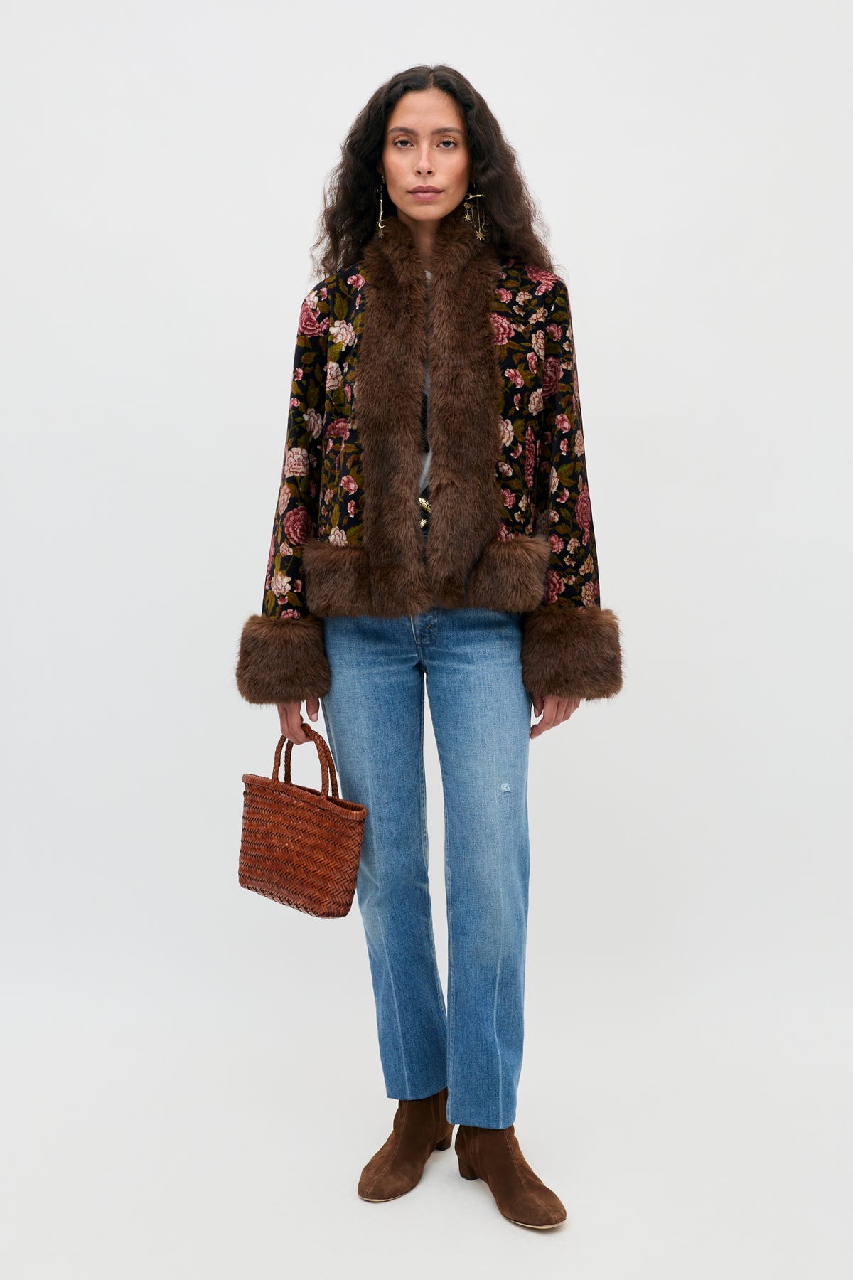 A woman stands facing forward, wearing the Jorgia - Enchanted Flora Green jacket with brown fur trim, blue jeans, brown boots, and holding a woven brown handbag. She has long wavy hair and a neutral expression against a plain background.