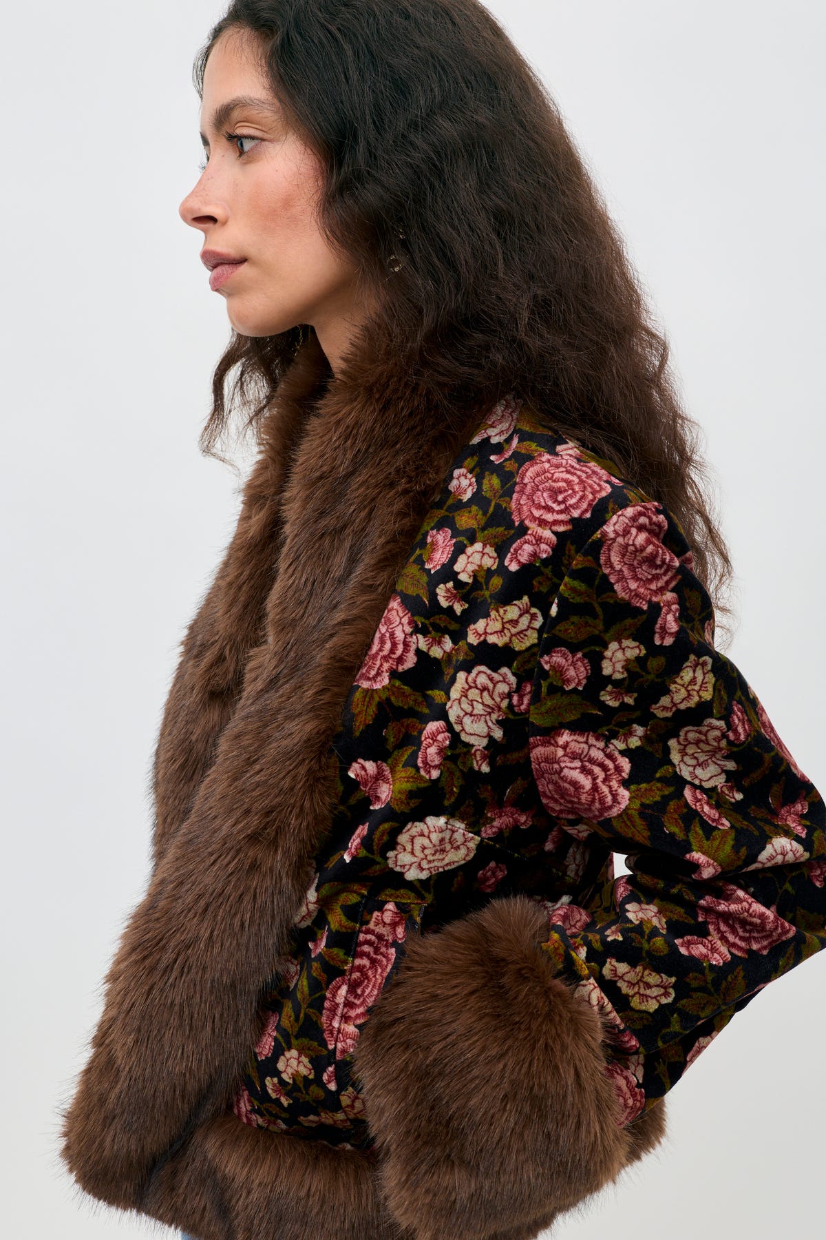 Woman in profile wearing the Jorgia - Enchanted Flora Green jacket, featuring a vibrant floral pattern and brown faux fur trim and cuffs, standing against a plain light background.
