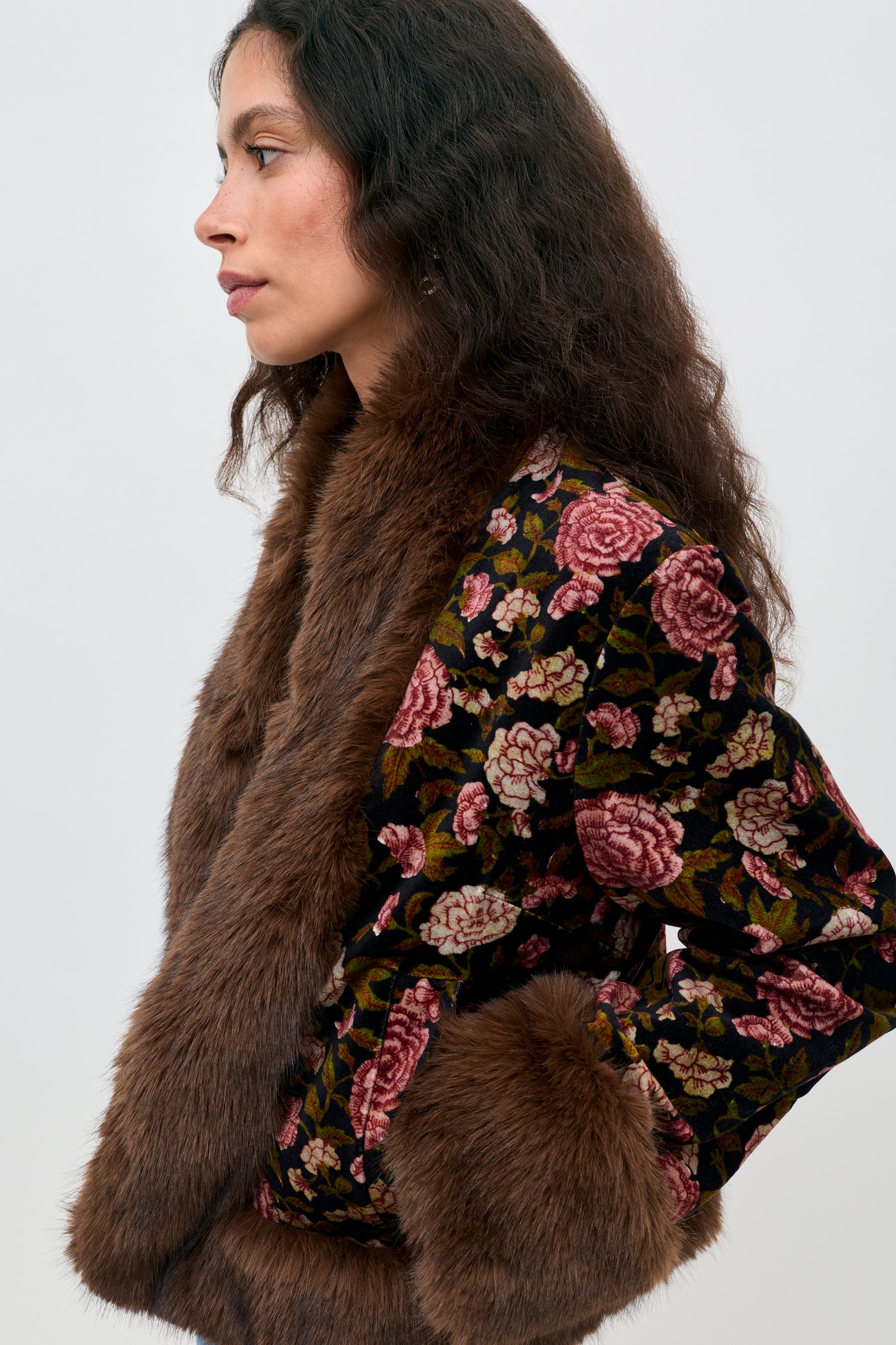 Woman in profile wearing the Jorgia - Enchanted Flora Green jacket, featuring a vibrant floral pattern and brown faux fur trim and cuffs, standing against a plain light background.