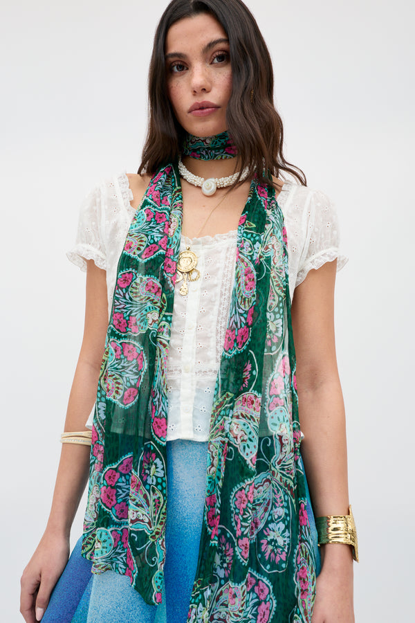 Wearing the Julien - Butterfly Bloom Green scarf, a woman pairs her white eyelet blouse and blue skirt with layered necklaces, a choker, gold bangles, and a ring, standing against a plain background.