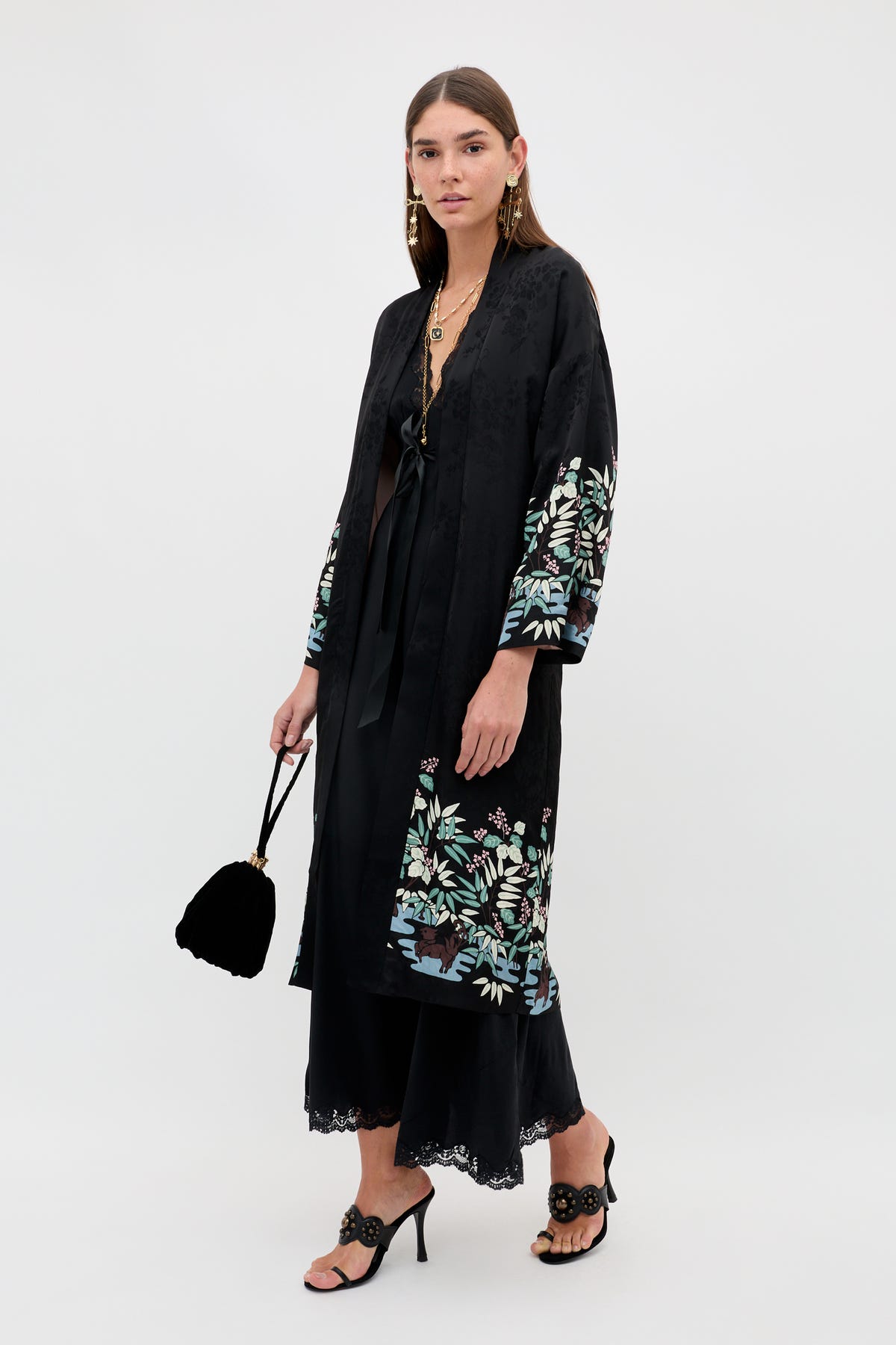 A woman models the Kaiden - Blossom Jungle Black, a long black kimono adorned with vibrant floral embroidery along the sleeves and hem, styled over a black dress with black heels and a small black handbag. She stands against a plain white background.