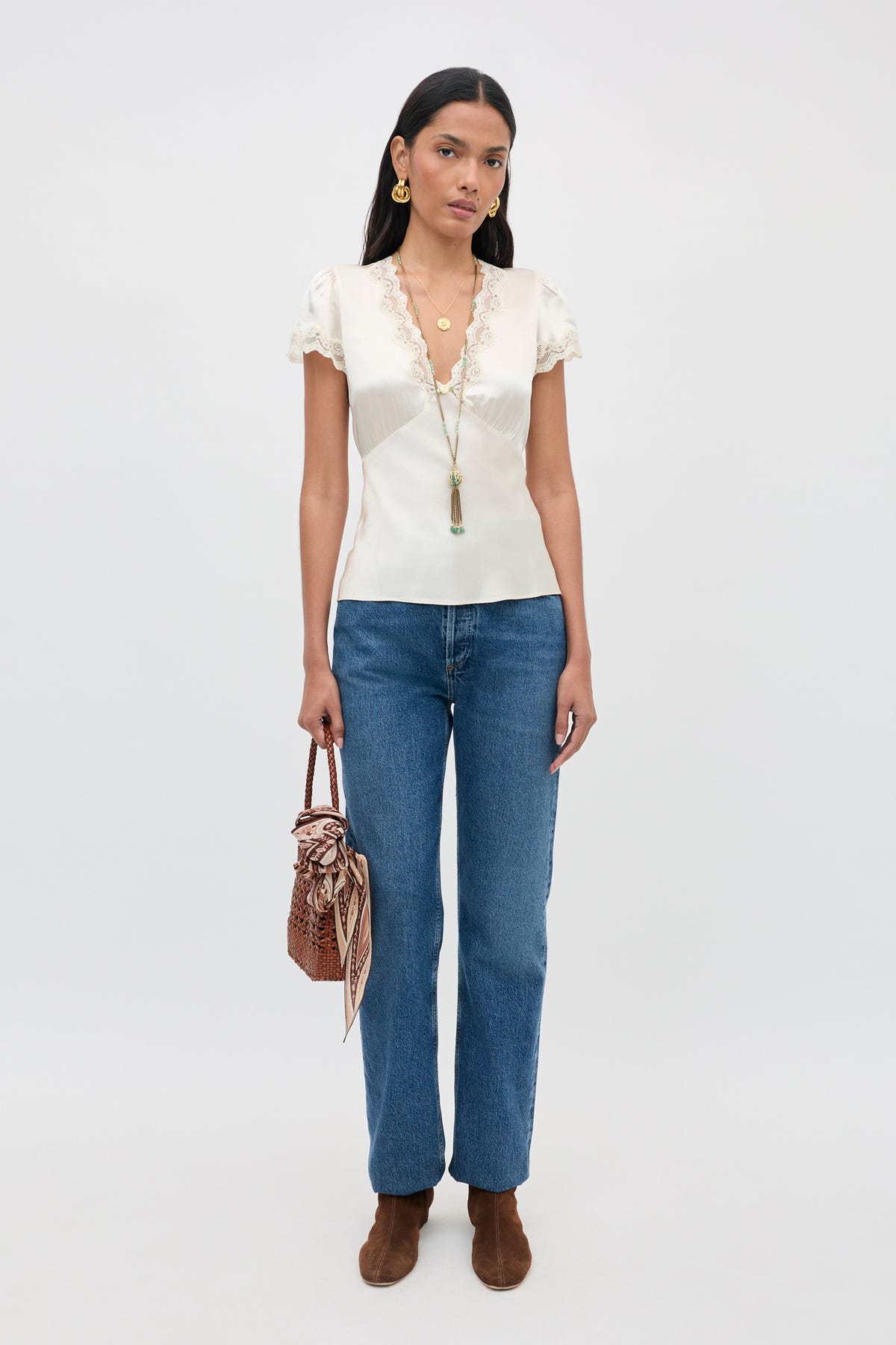Wearing the Karia - Champagne blouse, a woman pairs it with blue jeans, brown ankle boots, and carries a brown woven handbag. She accessorizes with gold jewelry and a long necklace, standing against a plain light-colored background.