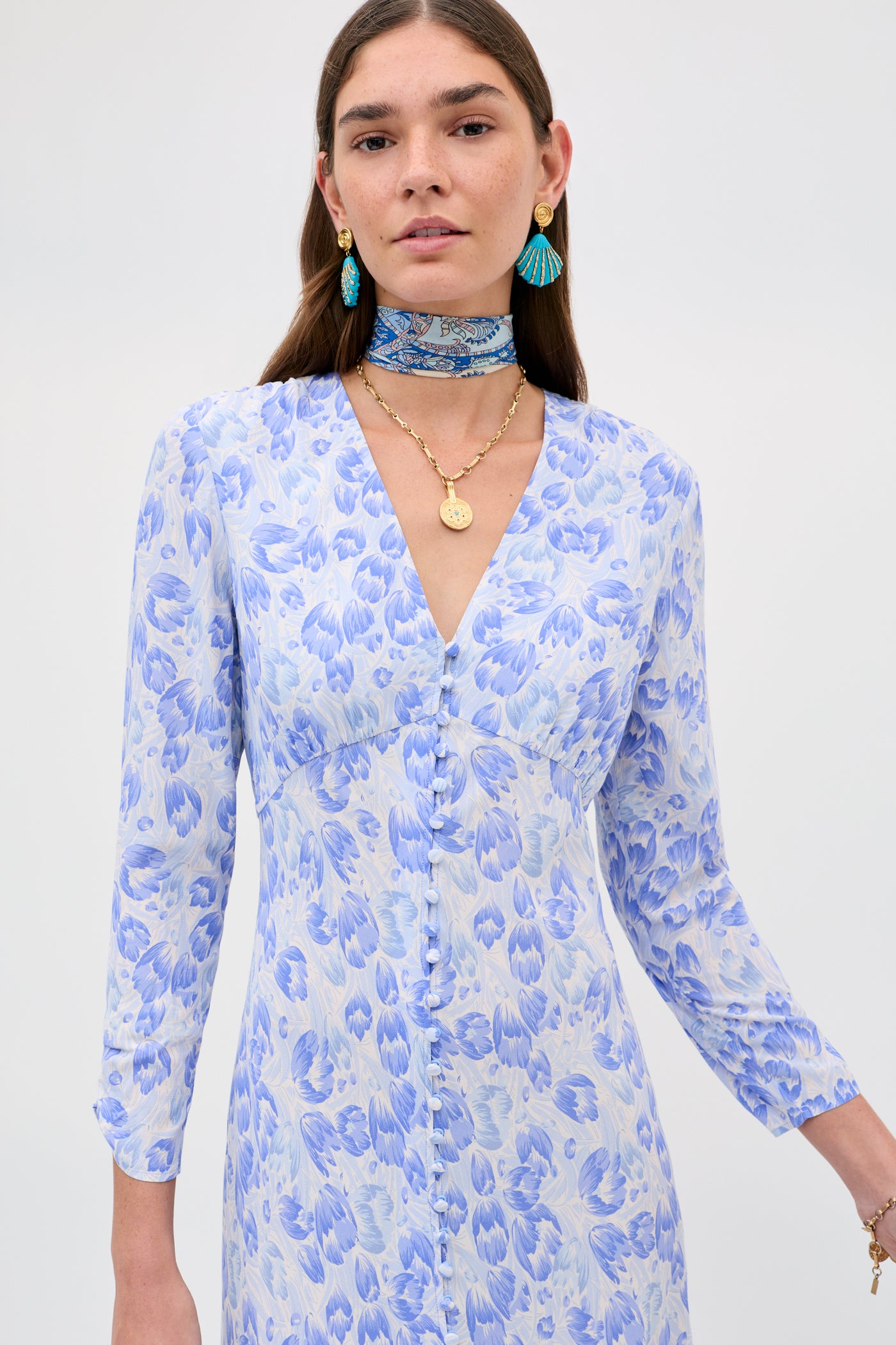 A woman wears the Katie - Tulip Fields Cornflower Blue dress, featuring long sleeves and a deep V-neckline—perfect for spring and summer—styled with layered gold necklaces, blue statement earrings, and a matching blue patterned choker.