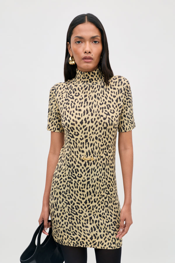 A person with long dark hair wears the Kylie - Miley Leopard Beige short-sleeve, high-neck dress and holds a black handbag. They accessorize with gold earrings and maintain a neutral expression while standing against a plain light background.