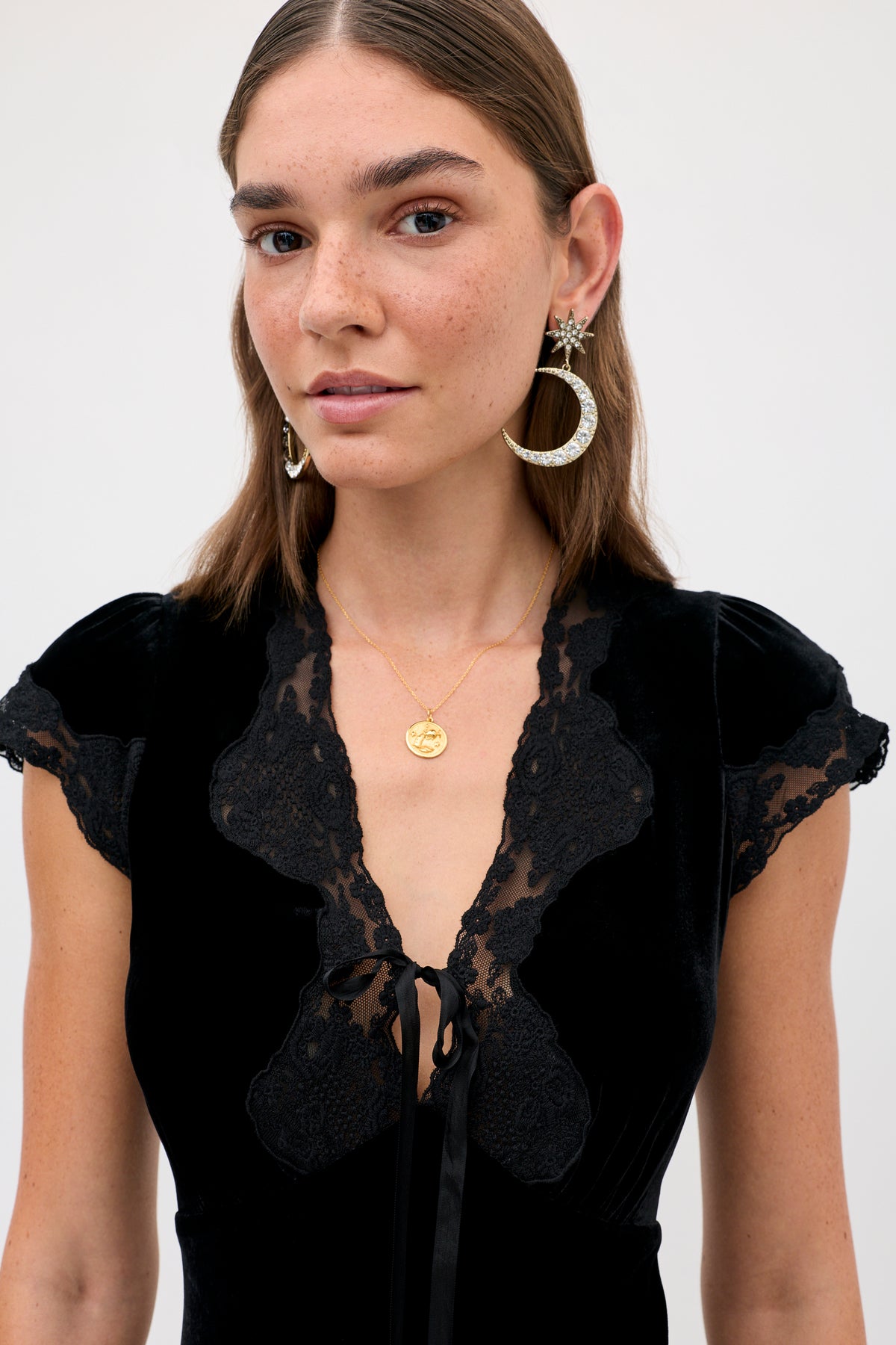 A woman wears the Lacie - Black velvet top with lace trim, a gold coin pendant necklace, and large crystal moon-and-star earrings.