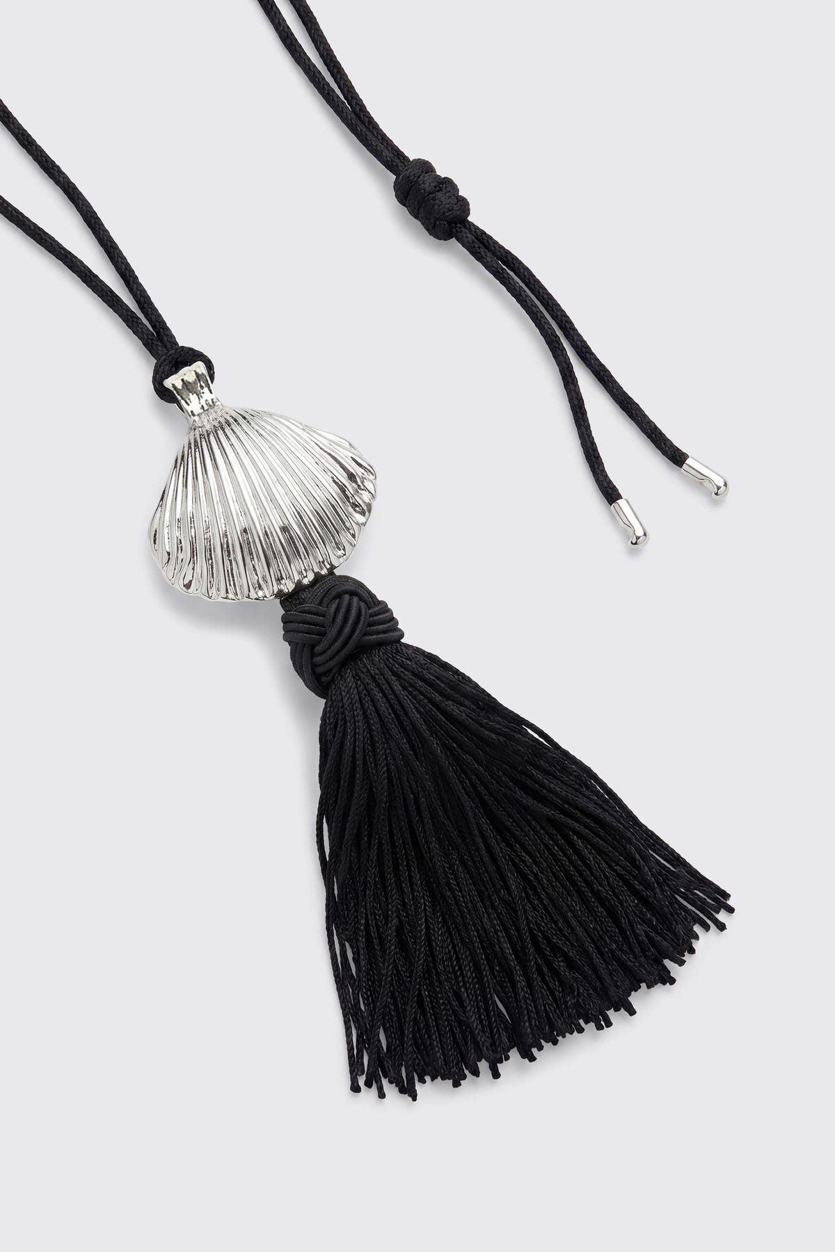 Liana - Silver: A necklace featuring a silver shell pendant and a black knotted tassel, suspended from a black cord with silver tips, displayed on a light gray background.