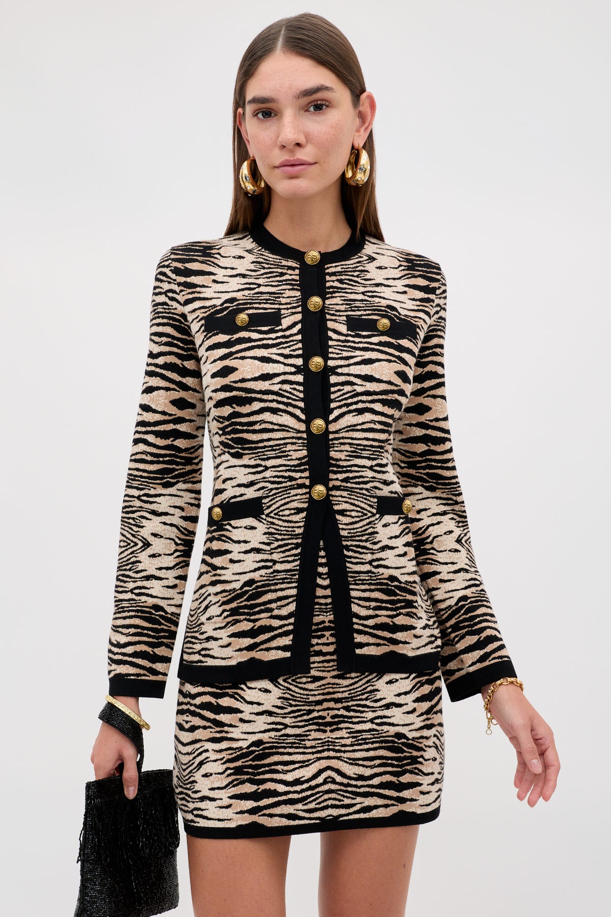 Wearing the “Liesel - Cooper Tiger Beige” set featuring a beige and black animal print jacket and skirt with gold buttons, she completes her look with gold hoop earrings and a black textured handbag while standing against a plain light background.