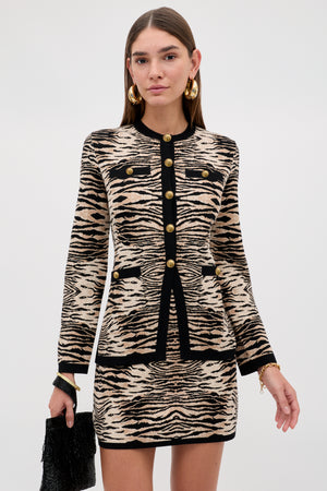 Wearing the “Liesel - Cooper Tiger Beige” set featuring a beige and black animal print jacket and skirt with gold buttons, she completes her look with gold hoop earrings and a black textured handbag while standing against a plain light background.