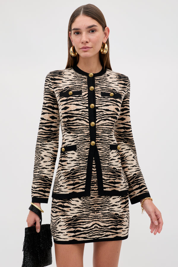 Wearing the “Liesel - Cooper Tiger Beige” set featuring a beige and black animal print jacket and skirt with gold buttons, she completes her look with gold hoop earrings and a black textured handbag while standing against a plain light background.