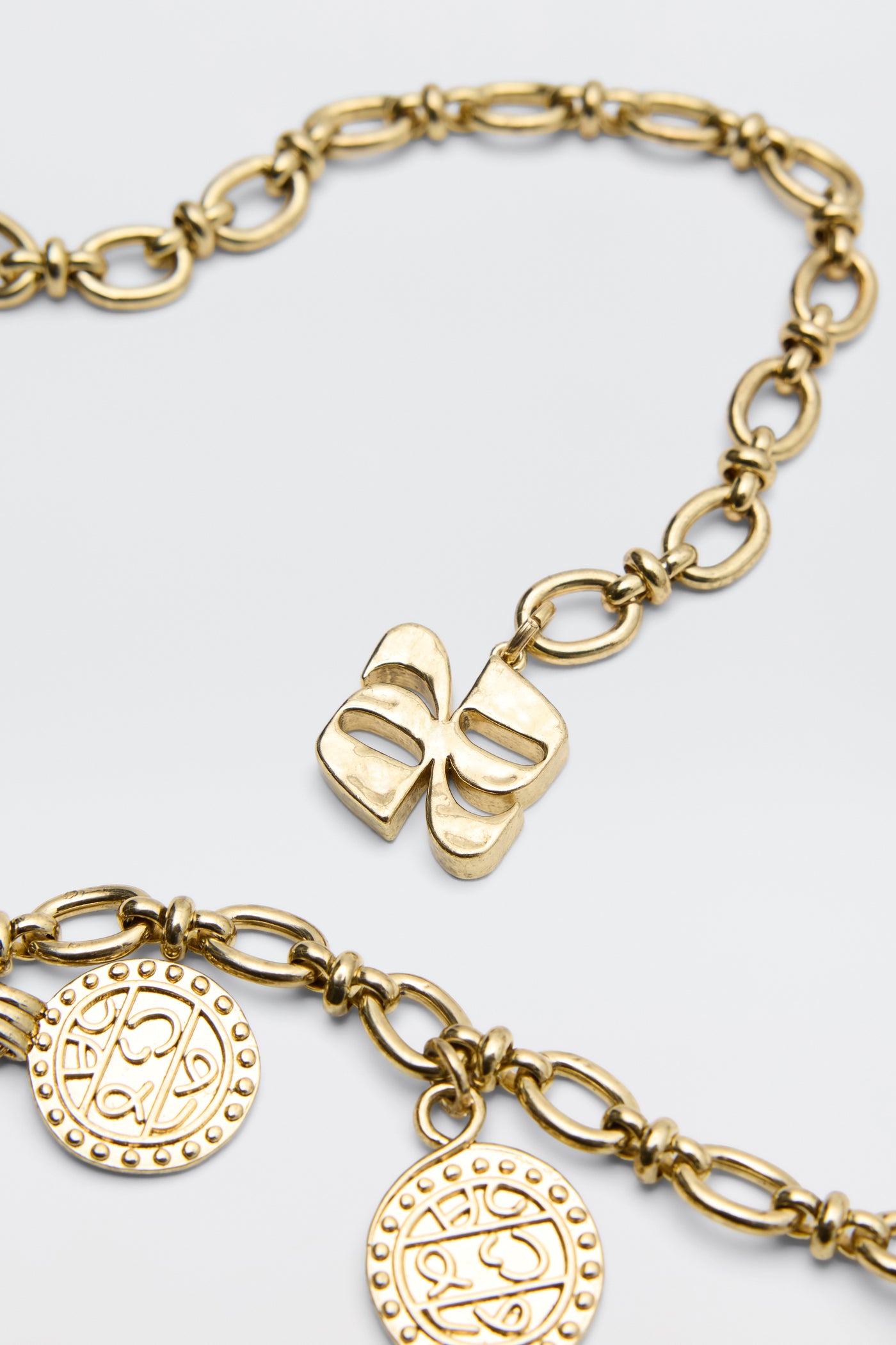 The Lita - Gold necklace features a bow-shaped charm and two round, engraved coin pendants on oval links, shown against a white background.