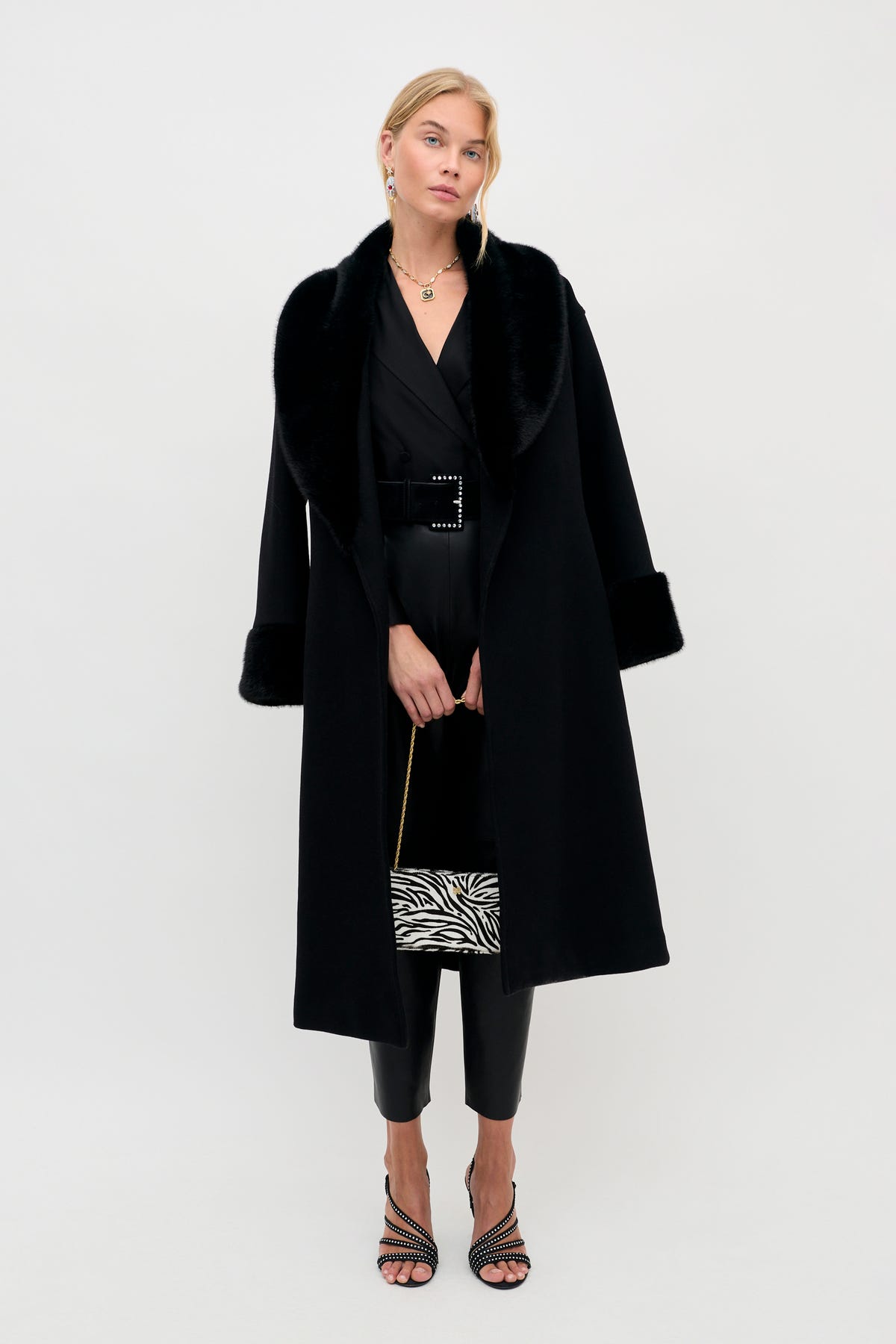 A woman wearing a Lorraine - Black coat with a faux fur collar and cuffs over a black belted outfit, holding a zebra-print handbag.