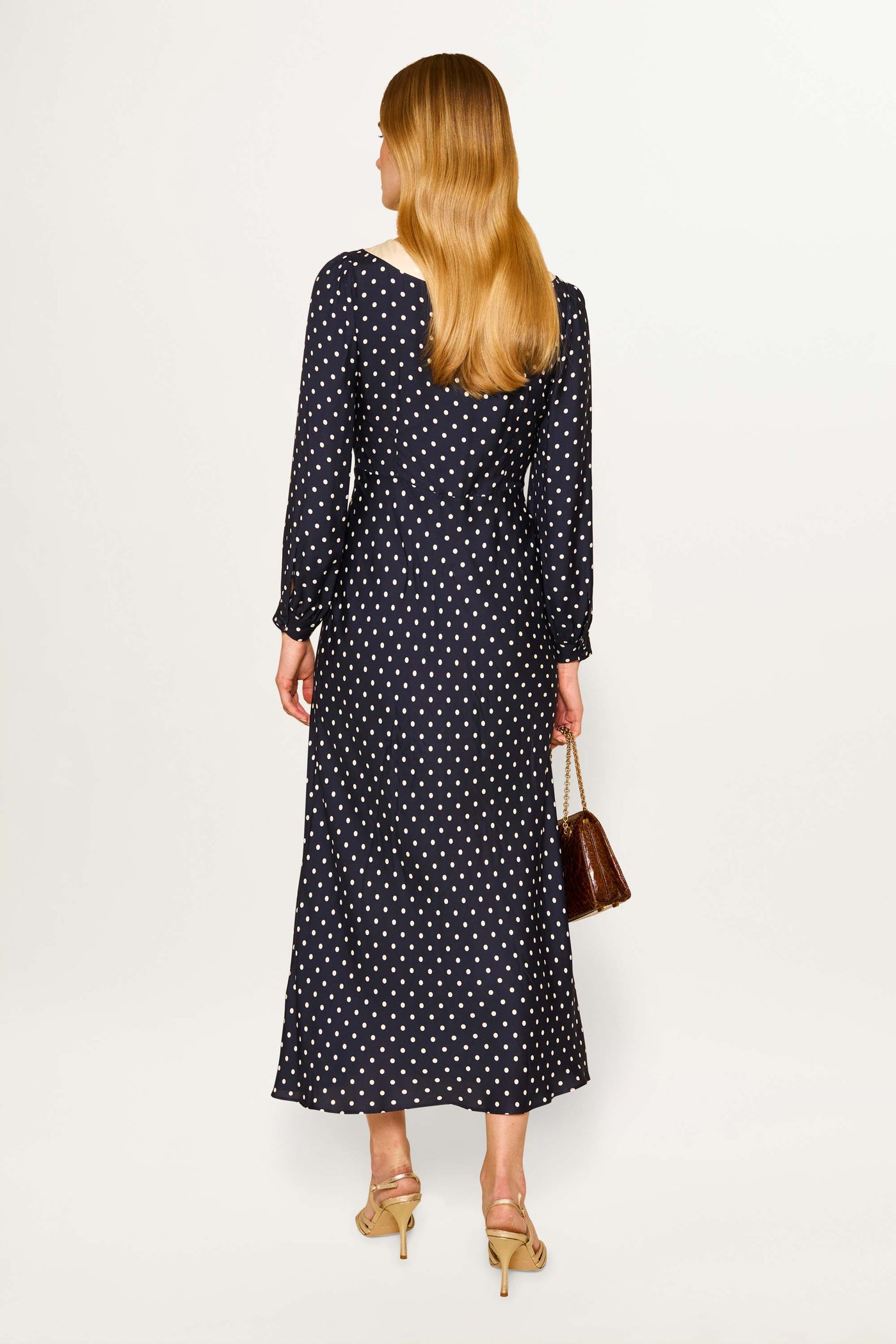 A woman with long blonde hair stands facing away, wearing the Lotta - Polka Dot Deep Navy dress with blouson sleeves. She carries a brown handbag and wears beige high-heeled sandals.