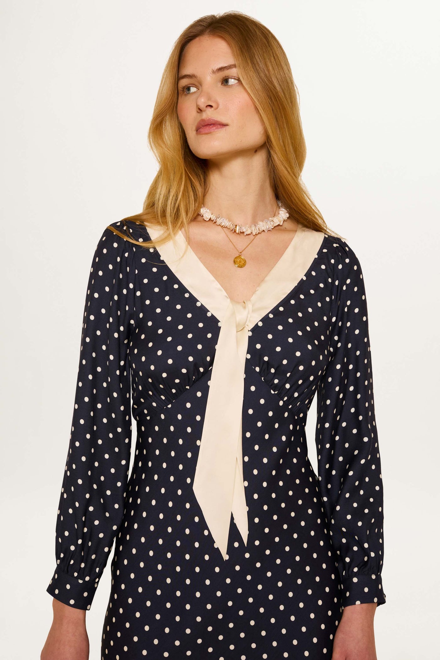 A woman with long blonde hair models the Lotta - Polka Dot Deep Navy dress, featuring white polka dots and a cream collar with bow, as she looks slightly to her left against a plain light background.