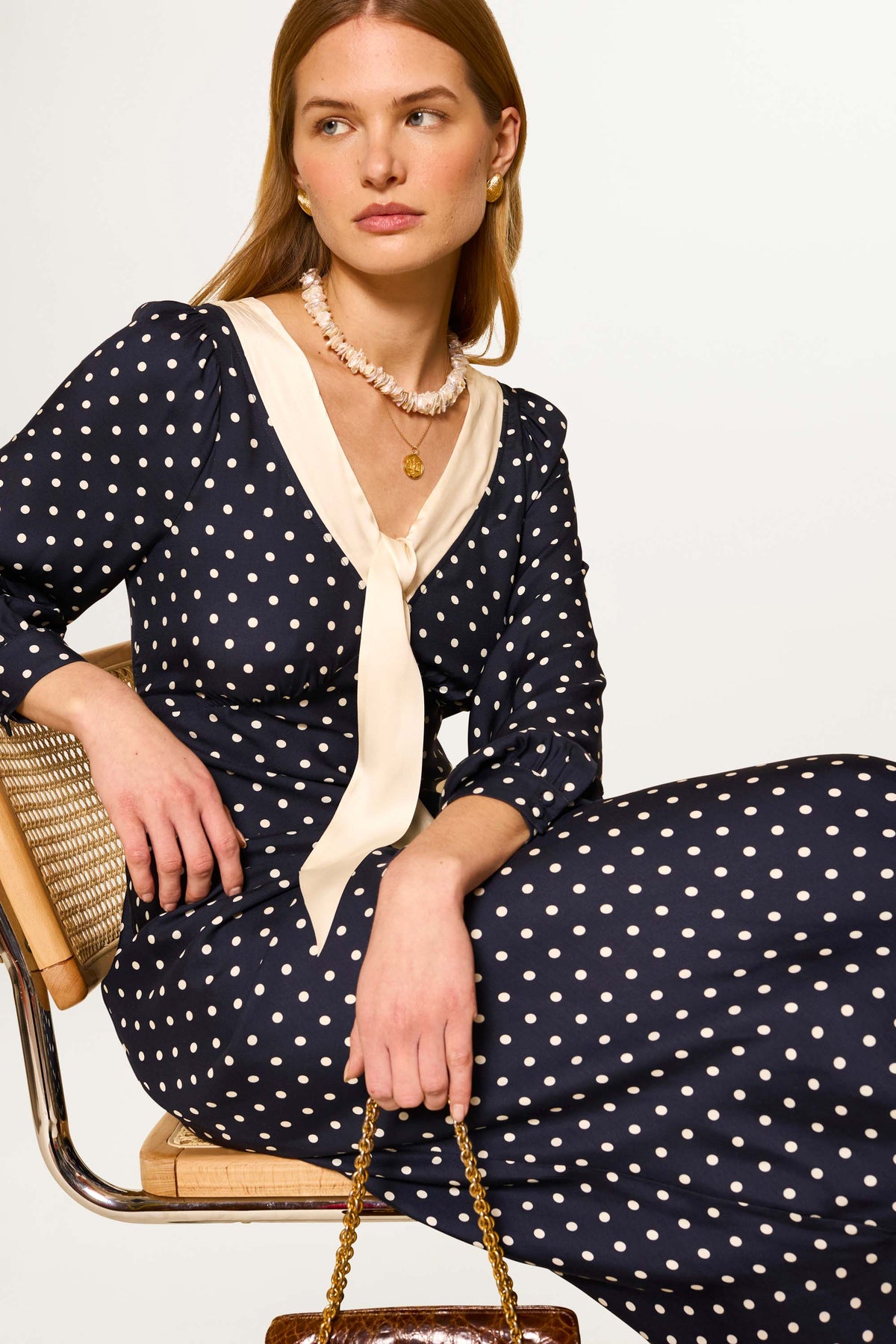 A woman in the Lotta - Polka Dot Deep Navy dress with a cream necktie sits on a chair, holding a textured handbag. She wears a pearl necklace, has light brown hair, and gazes to the side.
