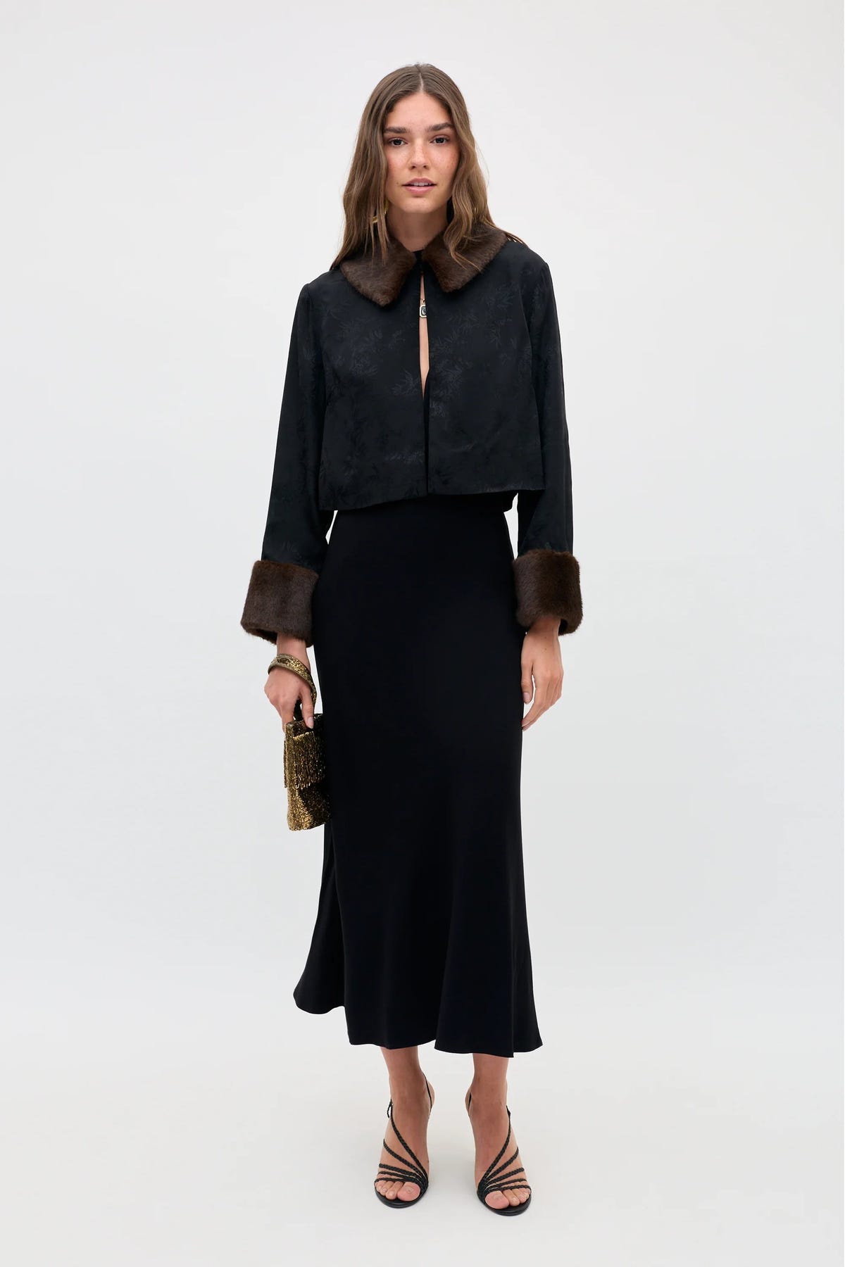 A woman stands facing forward against a plain light background, wearing the Luanne - Black long-sleeve jacket with faux fur collar and cuffs, a matching long black skirt, black strappy heels, and holding a small textured handbag.