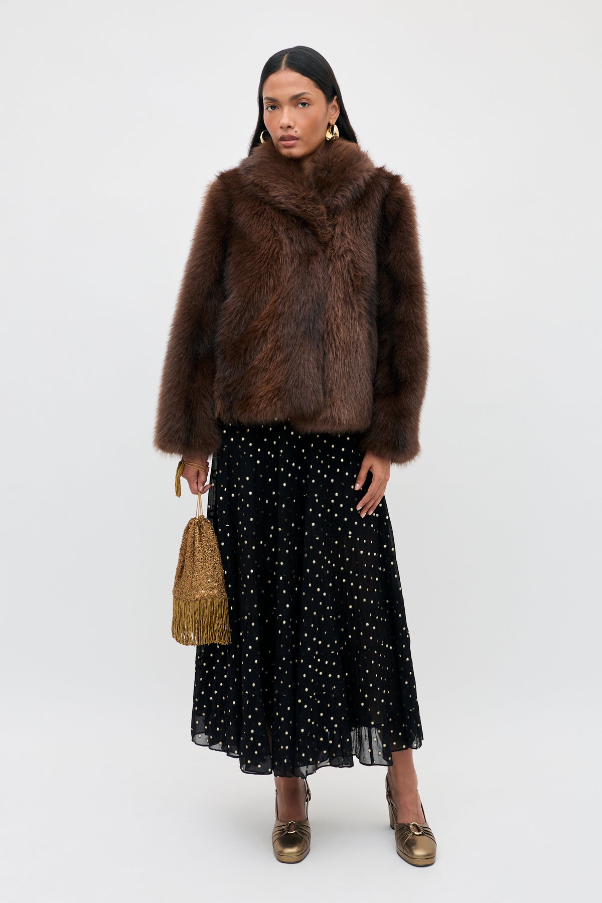 A person wearing a brown faux fur jacket and a Lucina - Gold Spot Black midi skirt, carrying a gold beaded handbag and wearing gold platform heels.