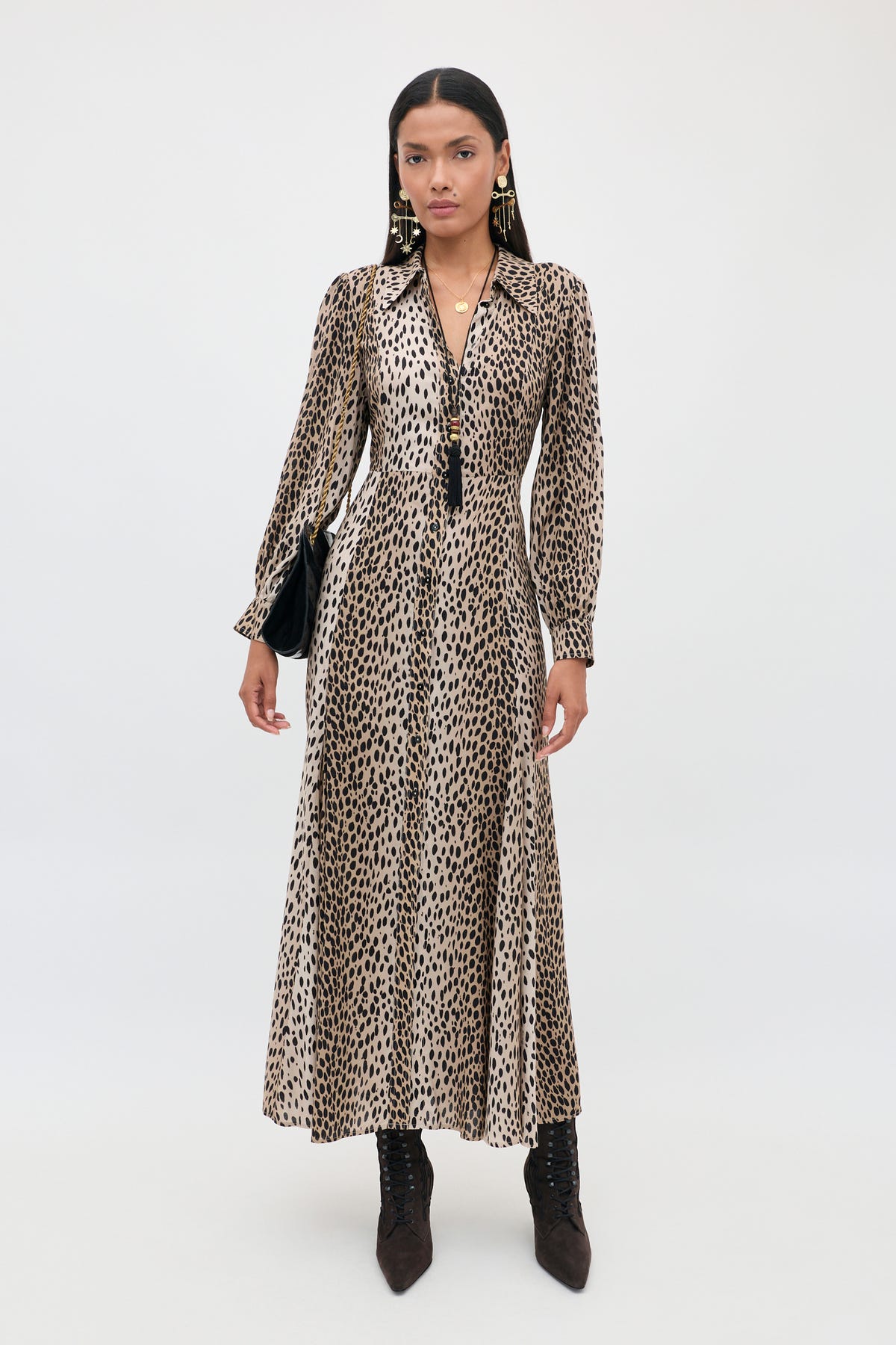 A woman stands against a plain light background wearing the Maddison - Antelope Spot Black, a long-sleeved, collared shirt dress featuring an antelope spot black print—styled with black boots, drop earrings, a necklace, and a black handbag—for the perfect petite-friendly everyday look.