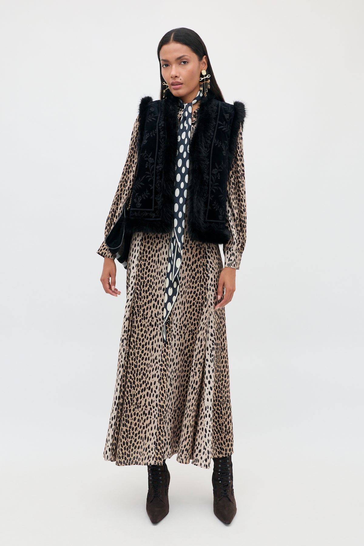 Petite woman standing against a plain background, wearing the Maddison - Antelope Spot Black long shirt dress paired with a black faux-fur vest, polka-dot scarf, statement earrings, and dark ankle boots. She has straight, dark hair and a neutral expression—embracing bold everyday styles.