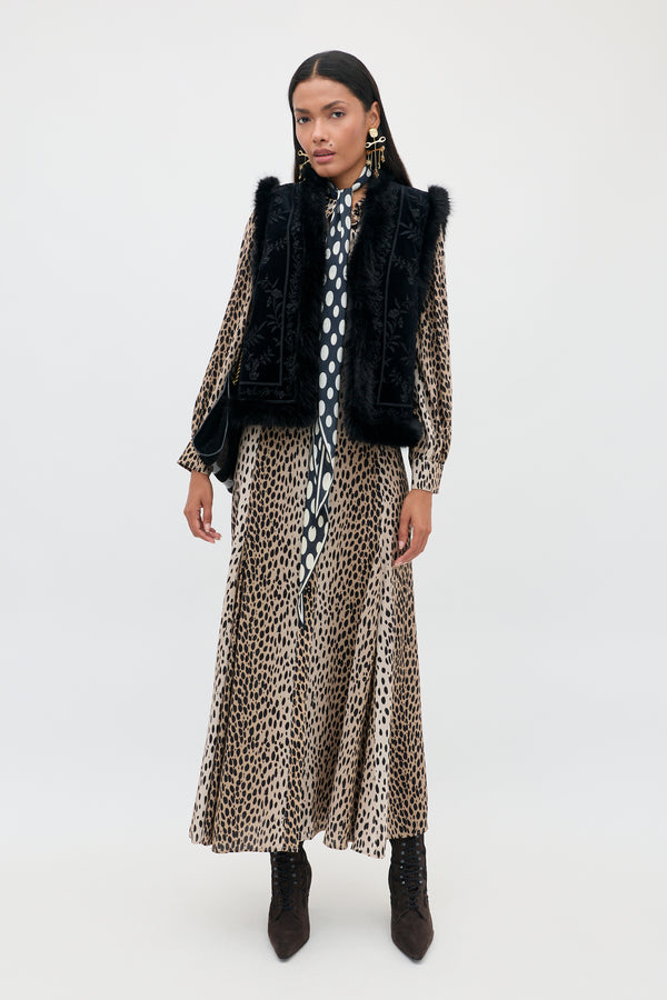 Petite woman standing against a plain background, wearing the Maddison - Antelope Spot Black long shirt dress paired with a black faux-fur vest, polka-dot scarf, statement earrings, and dark ankle boots. She has straight, dark hair and a neutral expression—embracing bold everyday styles.